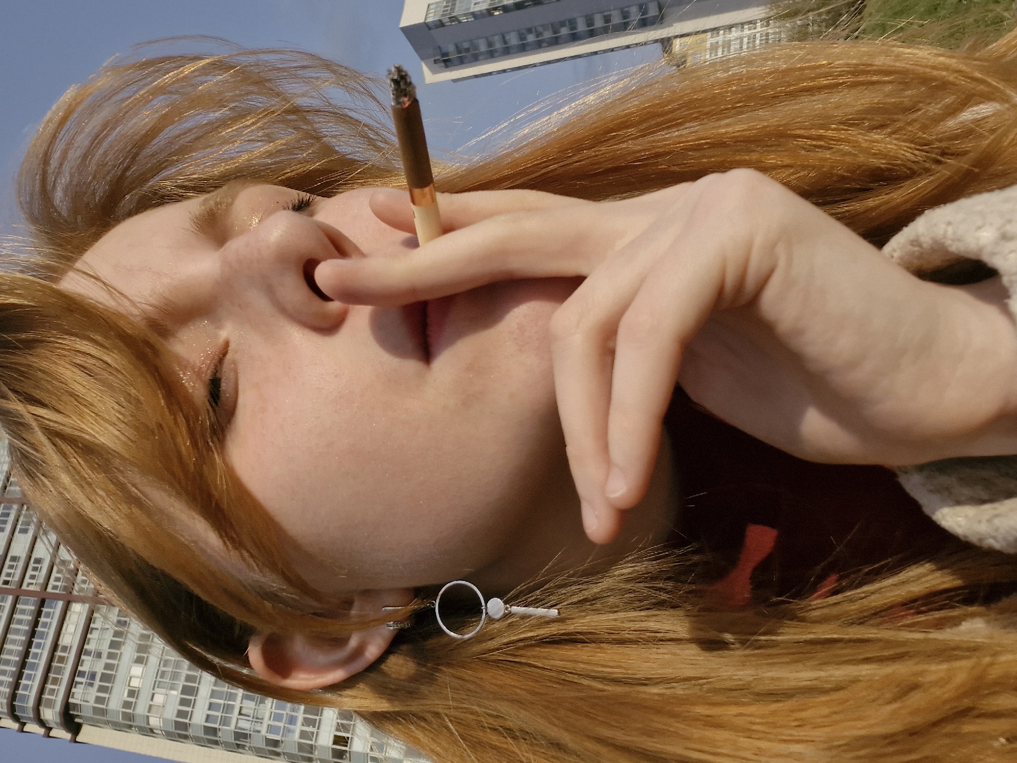 A close-up, Dutch-angle outdoor portrait features a young woman with fair skin, light freckles, and long, flowing reddish-blonde hair. She is depicted from a low perspective, looking upwards towards a clear blue sky, with her eyes closed or heavily squinted and her lips slightly parted. She is actively smoking a cigarette, which is held between her index and middle fingers with a visible ash tip, positioned near her mouth.
Her left ear is visible, adorned with a silver-colored earring that features a small circular hoop and a bar-like extension with a bead. A portion of a textured, light-colored garment, possibly a knitted sweater or jacket, is visible on her right side. The background reveals modern, multi-story urban buildings with numerous windows, suggesting a city environment, consistent with the context of Yekaterinburg, Russia. The bright, natural lighting indicates it is daytime, likely a sunny late morning or afternoon. The woman's relaxed posture and serene expression convey a sense of calm, contemplation, or enjoyment of the moment and the outdoor setting. There is no visible text in the image.