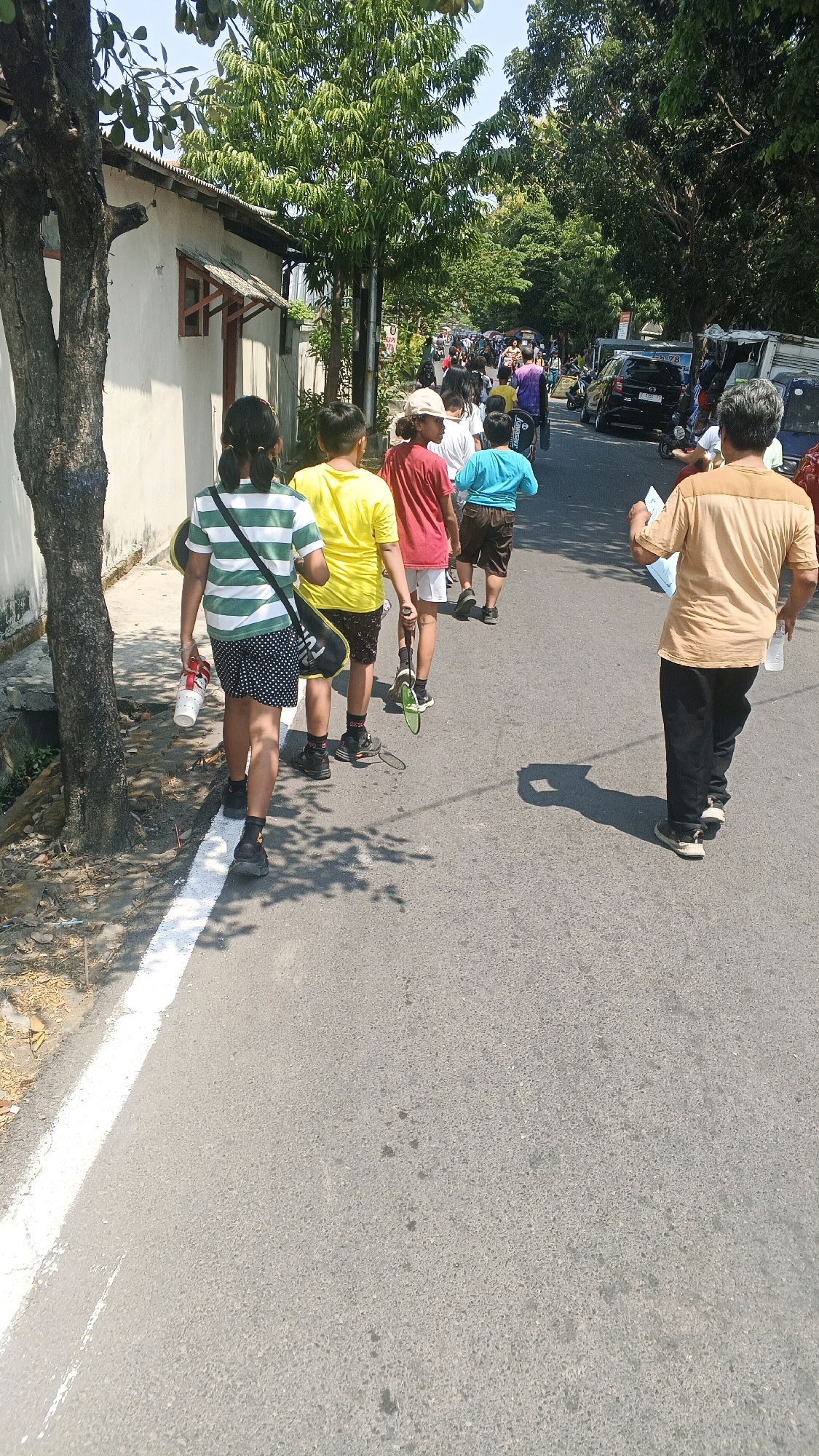 In Semarang, Indonesia, a group of children and adults are walking along a street. In the foreground, a young girl with braided hair wears a green and white striped shirt and black shorts with white polka dots. She carries a white cup and a black bag with a white logo. Behind her, a boy in a yellow t-shirt and black shorts walks with a badminton racket. Another boy in a red shirt and white shorts is also walking, holding a badminton racket. Further back, a boy in a blue shirt and brown shorts joins the group. An adult man in a tan shirt and black pants walks away from the viewer, holding a white bottle and a piece of paper. Several other people and vehicles, including cars and motorcycles, are visible further down the street, suggesting a bustling urban environment. The scene is illuminated by bright sunlight, casting shadows on the asphalt road. The background features trees and buildings, indicating an outdoor setting in a city.