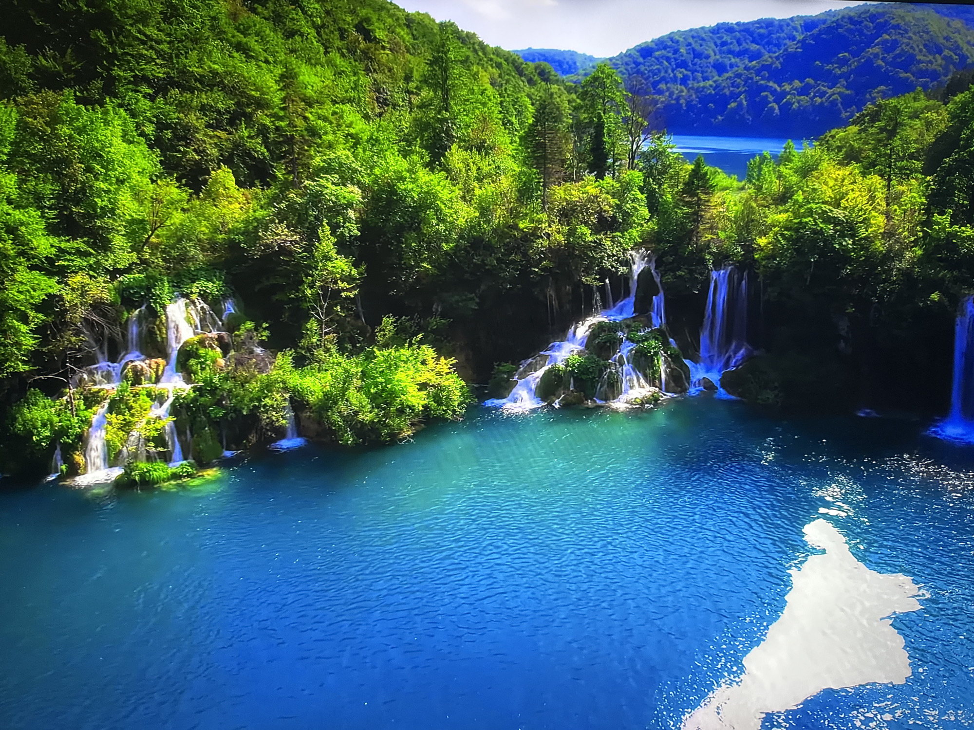 The image shows a scenic landscape with a vibrant blue lake or river, surrounded by lush green trees and several waterfalls cascading down rocky formations. In the distance, rolling hills covered in dense vegetation create a layered background. The water is clear and reflects the surrounding greenery and the sky, which appears partly cloudy. The overall impression is one of natural beauty and tranquility. This image does not depict any location in Burgas, Bulgaria.