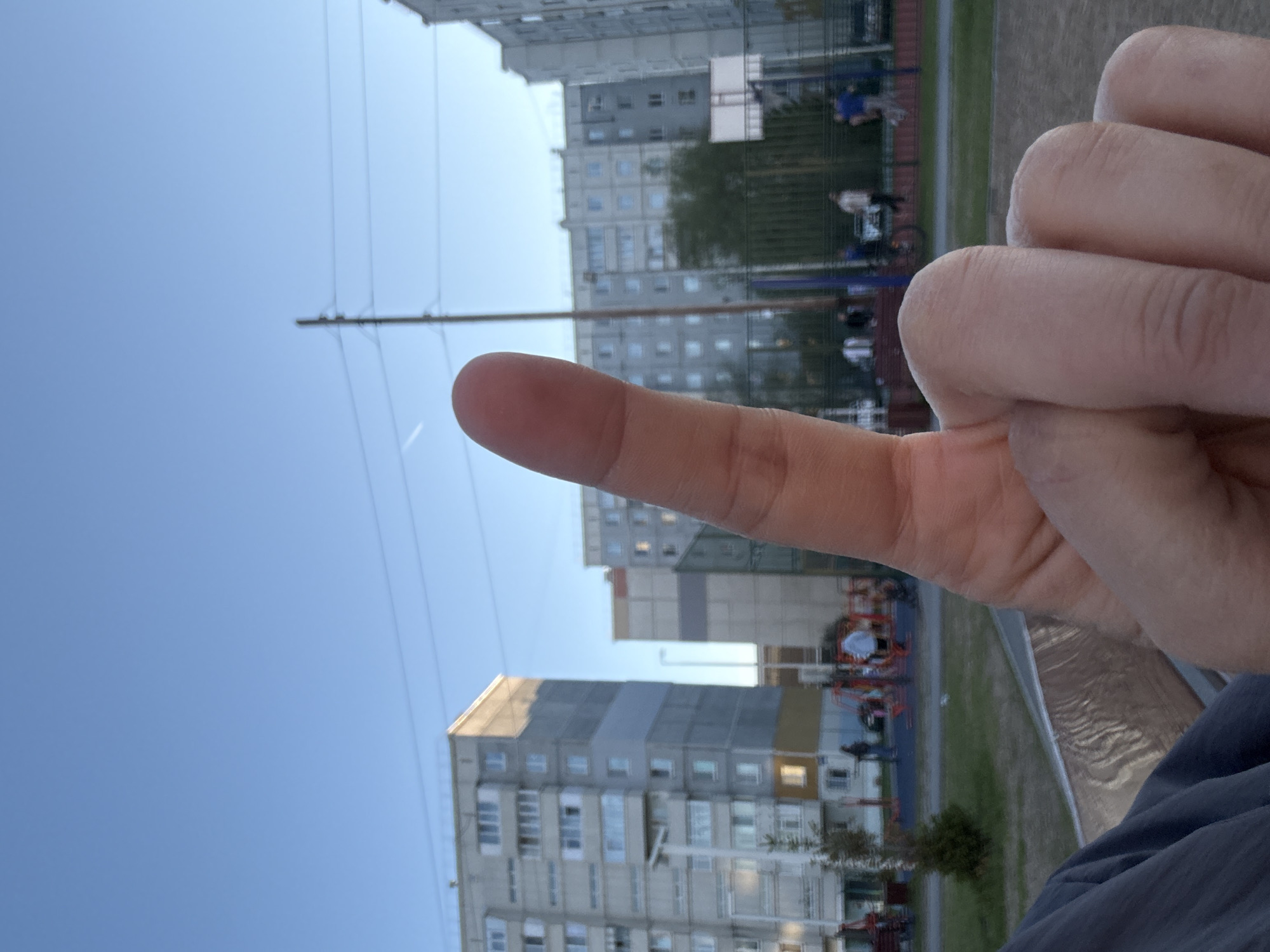 A close-up, vertically oriented photograph depicts a hand with a finger extended upward against an urban backdrop, likely taken in Tobolsk, Russia. The foreground features a person's hand, obscuring the lower right portion of the frame. The extended finger points toward the sky.
In the background, several multi-story apartment buildings are visible. A playground area with red and orange play structures is present between the buildings. There are small groups of people present on the playground, but they are far enough to where details can't be determined. Overhead power lines run across the sky. The sky is blue, suggesting the photograph was taken during daylight hours.