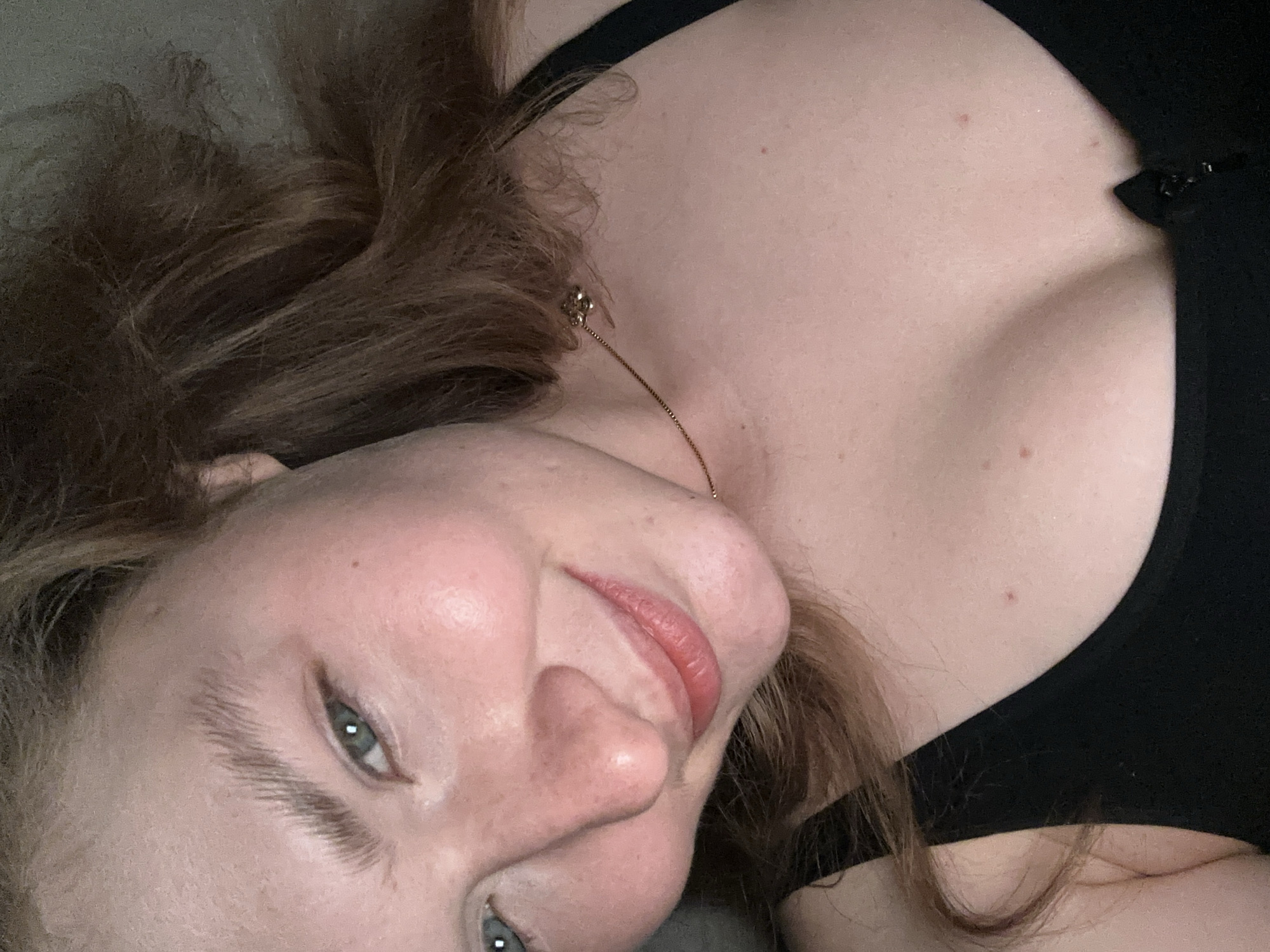 A close-up, tilted portrait shows a fair-skinned person with light brown hair and blue eyes. They are wearing a black top and a delicate gold necklace. The background is a plain, light-colored surface. The person is smiling slightly. The location is not discernible from the image.