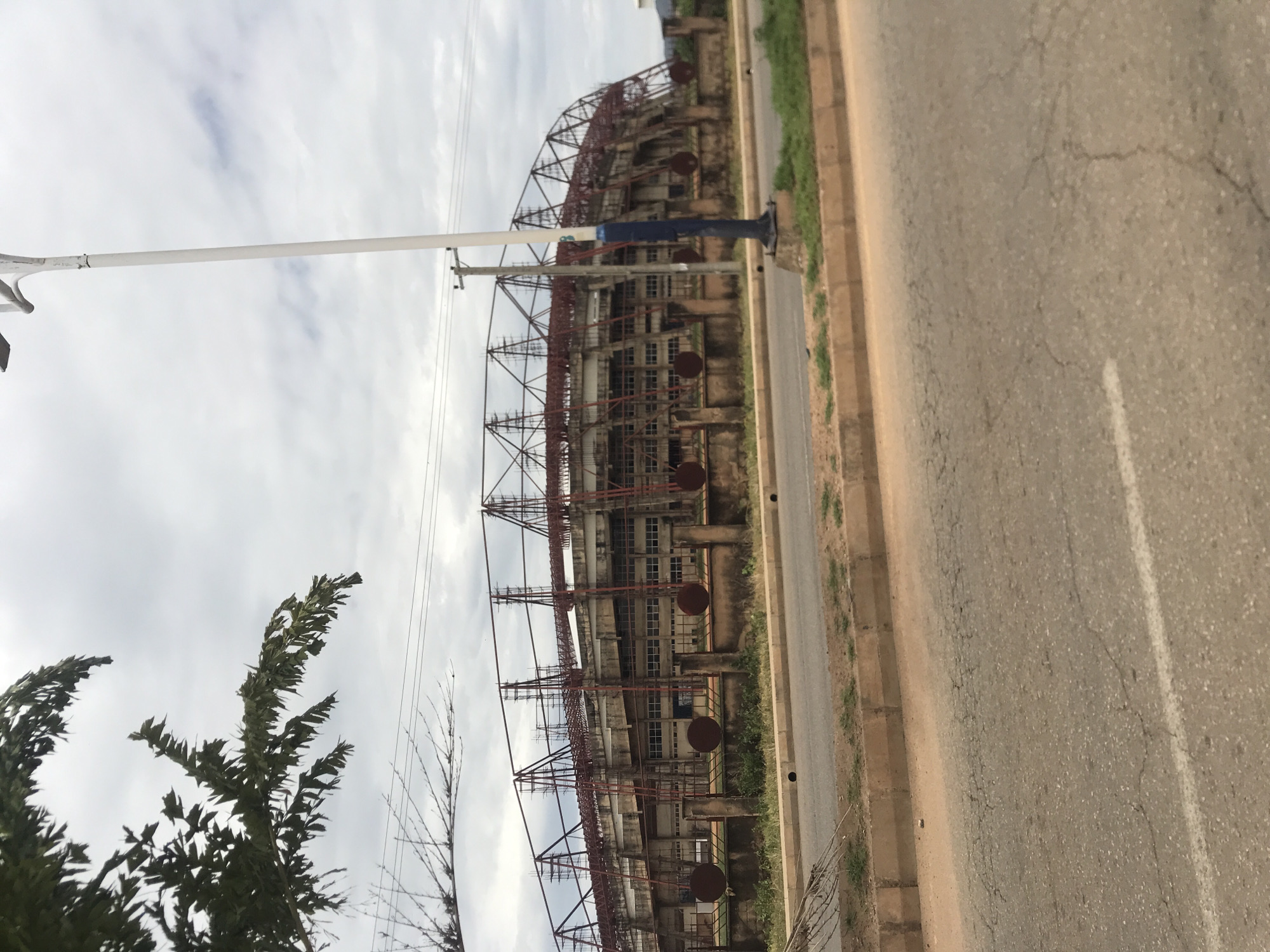 The image presents an exterior view of a large stadium structure, located in New Stadium, Nigeria, under an overcast sky. The stadium's main body is constructed of concrete, featuring multiple visible levels with numerous rectangular window-like openings. Above the concrete structure, an extensive, exposed framework of red metal trusses forms the upper part of the stadium, suggesting an ongoing construction or renovation phase for a roof. A low perimeter wall or fence stands in front of the stadium, characterized by several large, circular, rusted metal discs attached at regular intervals.
In the immediate foreground, an asphalt road with visible cracks and a faint white line runs horizontally across the bottom of the frame, separated by a concrete curb from a strip of dirt and sparse green vegetation. To the left, dark green foliage from a tree partially enters the upper left corner, with more sparse, bare branches extending from the lower left towards the mid-ground. On the right side, a tall, slender street light pole, white with a dark blue lower section, extends vertically from the foreground into the upper frame. Behind this, a wooden utility pole supports multiple overhead power lines that traverse the mid-ground horizontally. The sky is uniformly gray and cloudy. No people or vehicles are visible in the scene.