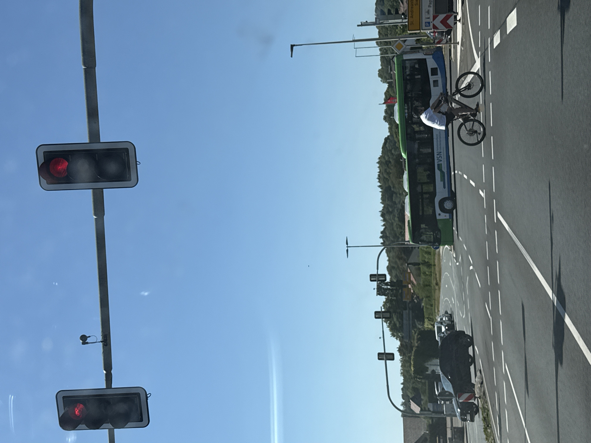 The image shows a street intersection with traffic lights, cars, a bus, and a cyclist. The traffic lights are red. A black car is stopped at the intersection, and a white car is behind it. A green bus is turning at the intersection. A cyclist is riding across the intersection. The scene appears to be in Herzberg, Germany, as suggested by a sign for "St. Andreasberg". The sky is blue and clear, and the sun is shining.