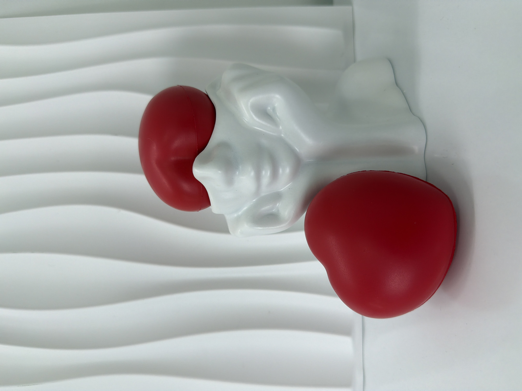 The image displays a white, wavy background and a white surface in the foreground. On the surface, there are two red heart-shaped objects and a white, sculpted object resembling a face. One heart is positioned to the left of the face sculpture, and the other is placed on top of the sculpture, covering its eyes. The face sculpture has its hands resting on its cheeks. The image is a still life composition with no discernible location in Bac Nha Trang Ward, Vietnam.