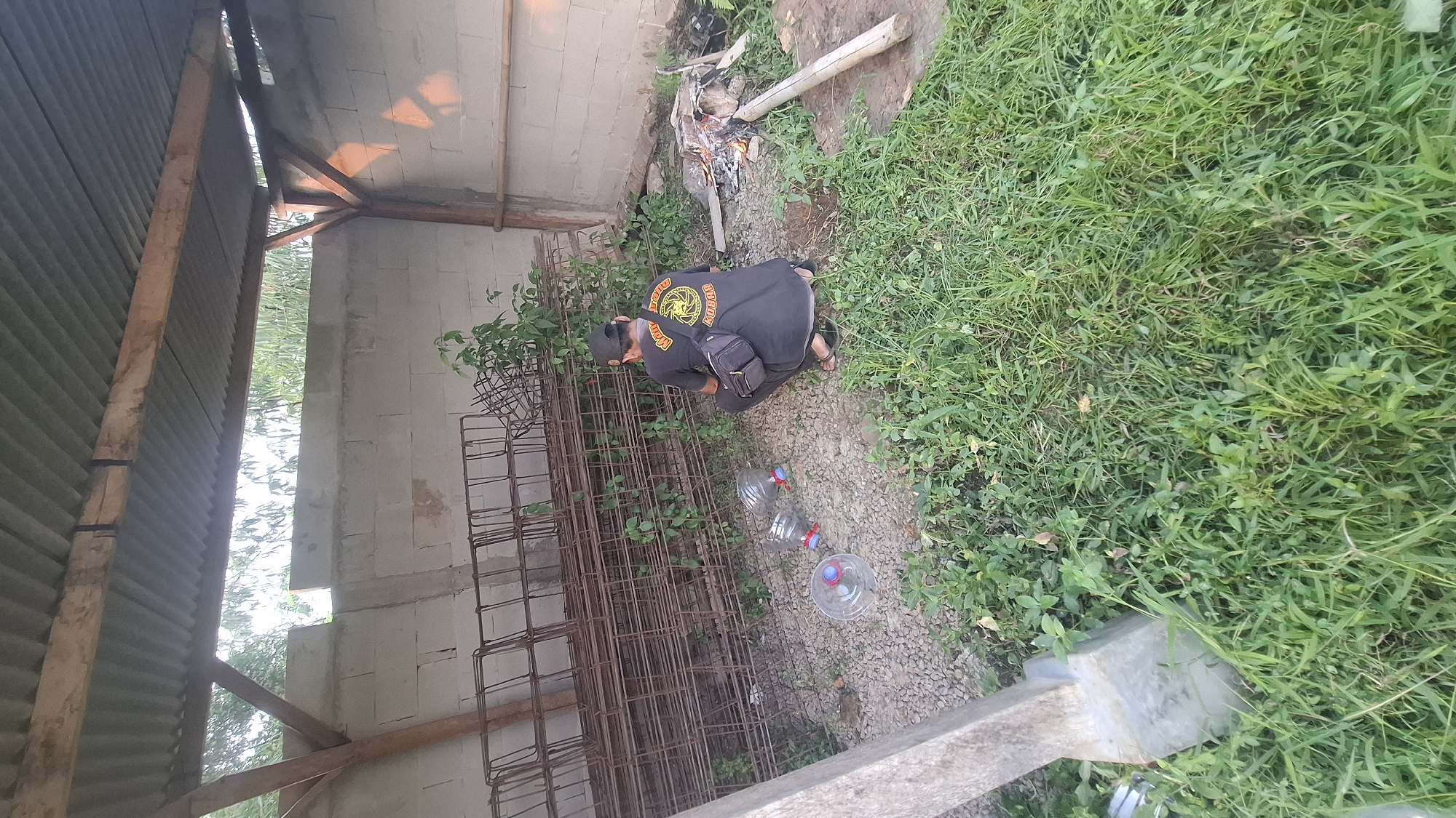 In Rahong, Indonesia, a man in a black t-shirt and cap is squatting near a pile of rebar. He has a black fanny pack across his chest. In front of him are three plastic bottles with red caps. To his right, a small fire burns in a pit with a long piece of wood next to it. The scene appears to be in an outdoor, possibly rural, area with a corrugated metal roof supported by wooden beams overhead. The ground is covered in gravel and grass, and there is a concrete pillar to the left of the frame. The wall behind the man is made of cinder blocks.