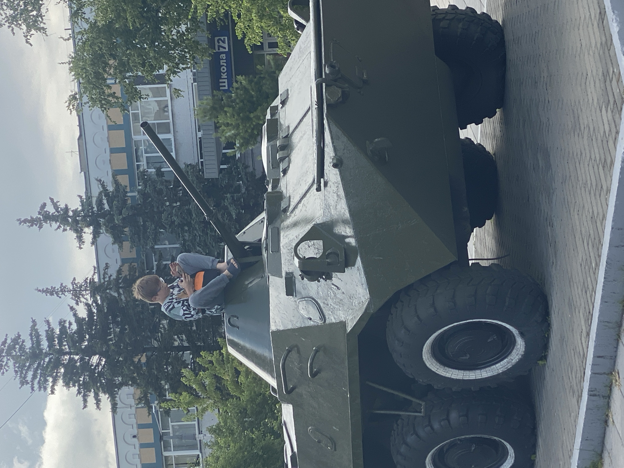 A young boy wearing glasses and a patterned shirt sits on top of a green armored personnel carrier, looking at his phone. He has an orange backpack or bag next to him. The APC is parked on a paved surface with a gray curb in the foreground. In the background, there is a building with the sign "Школа 72" (School 72) and several tall evergreen trees. The sky is overcast. The image appears to have been taken in Novokuznetsk, Russia.