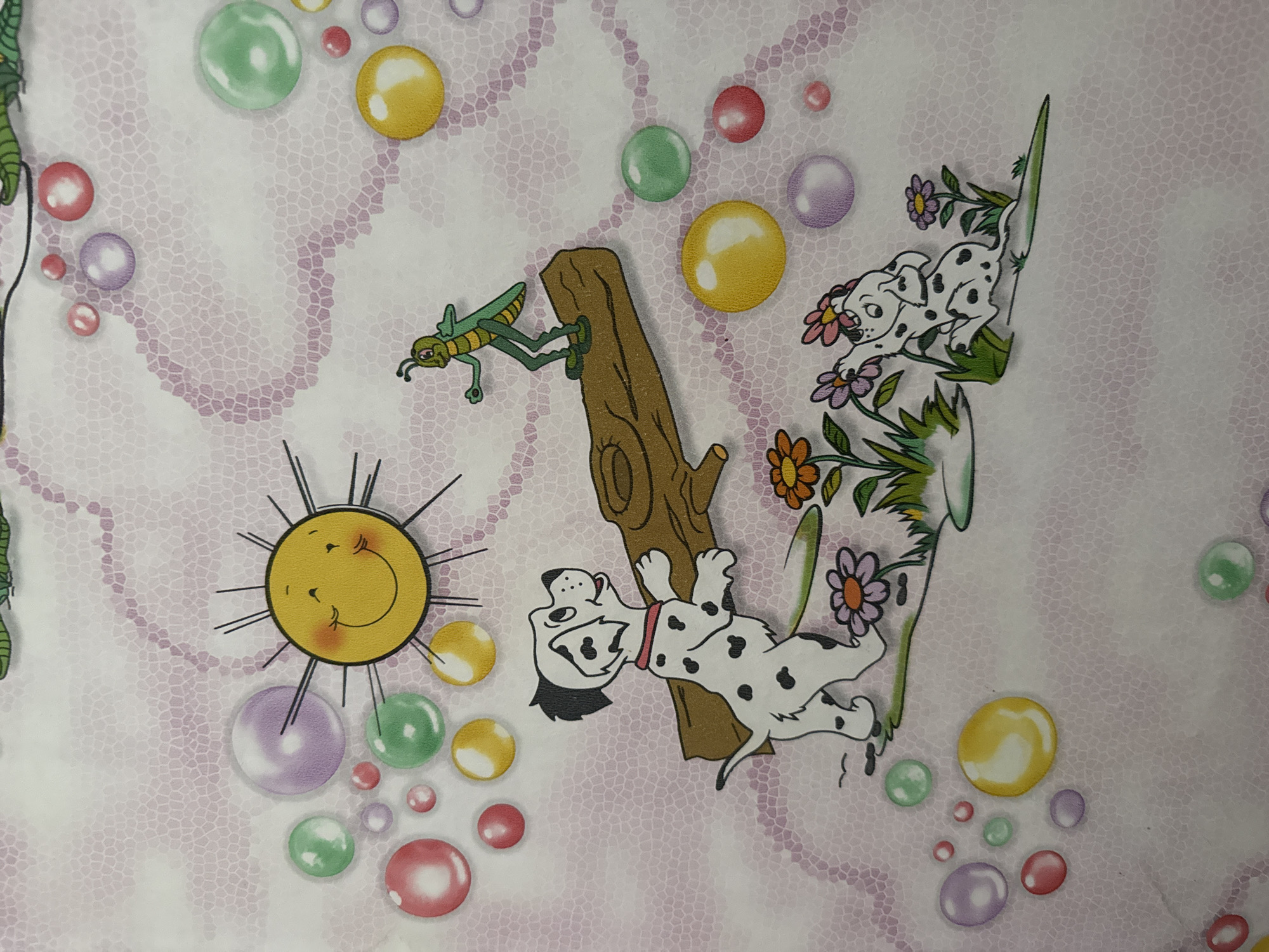The image depicts a cheerful cartoon scene. A smiling sun with rays is positioned in the upper left quadrant. Below and to the right of the sun, a green grasshopper stands on a brown log. In the lower portion of the image, two Dalmatian dogs are present. One dog, on the left, is white with black spots and a red collar, looking upwards and to the right. The other Dalmatian, on the right, is also white with black spots and appears to be playfully reaching towards flowers. The background features numerous colorful, translucent bubbles in shades of pink, purple, yellow, and green, scattered across a light pink, textured surface resembling a mosaic. This scene is a design on fabric, possibly for children's bedding or clothing. The location in the city of Bălți, Moldova, is not discernible from the image itself.