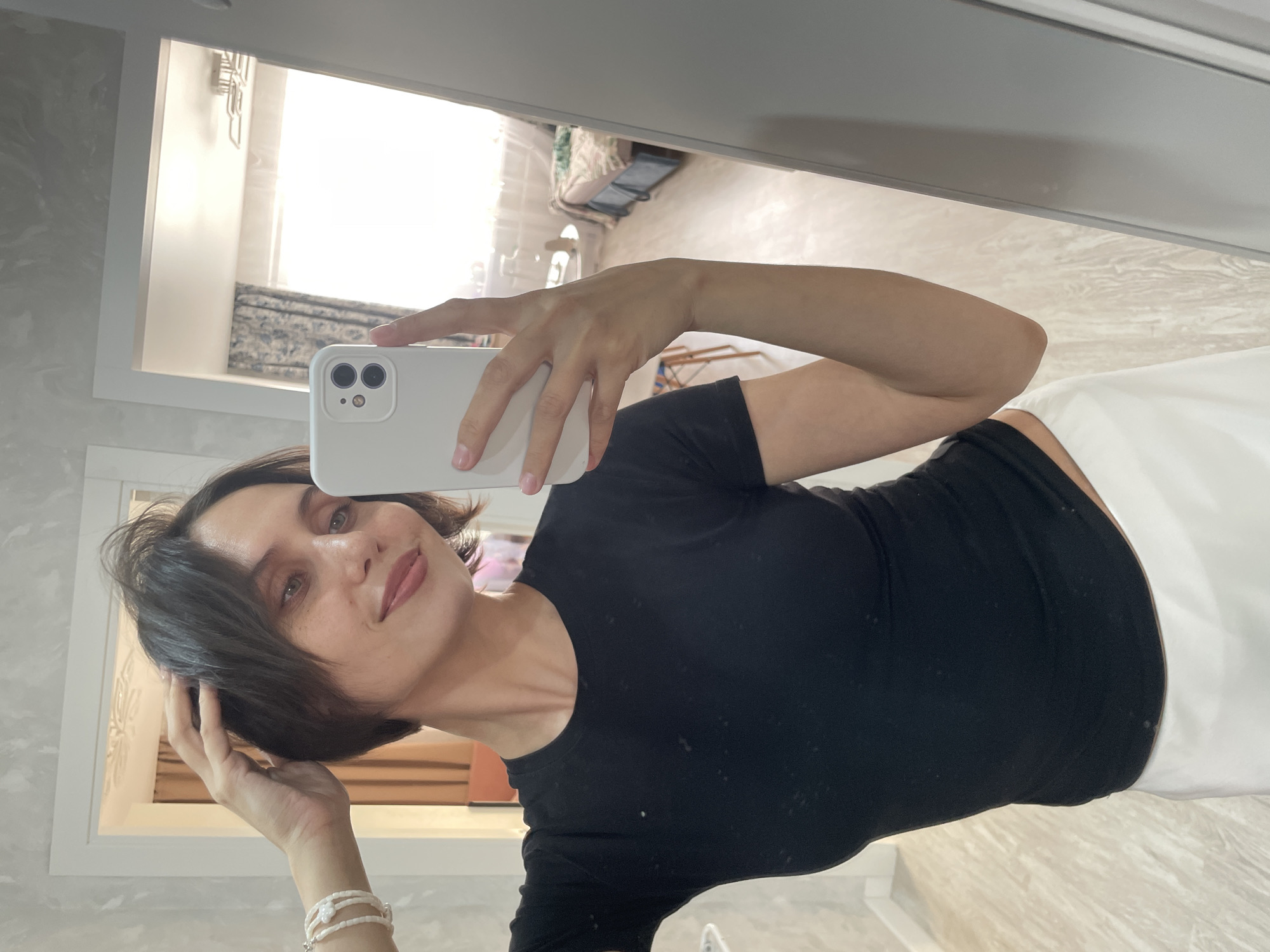 A woman is taking a selfie in a mirror. She is wearing a black t-shirt and a white garment (possibly a towel). She is holding a white smartphone with multiple cameras in her right hand. She is touching her short, dark hair with her left hand. She is also wearing a multi-strand bracelet on her left wrist. The reflection in the mirror reveals a bright interior space with a window and what appears to be a glimpse of furniture (possibly a bed or sofa). The location, Izhevsk, Russia, is mentioned without any visual confirmation, but is to be included.