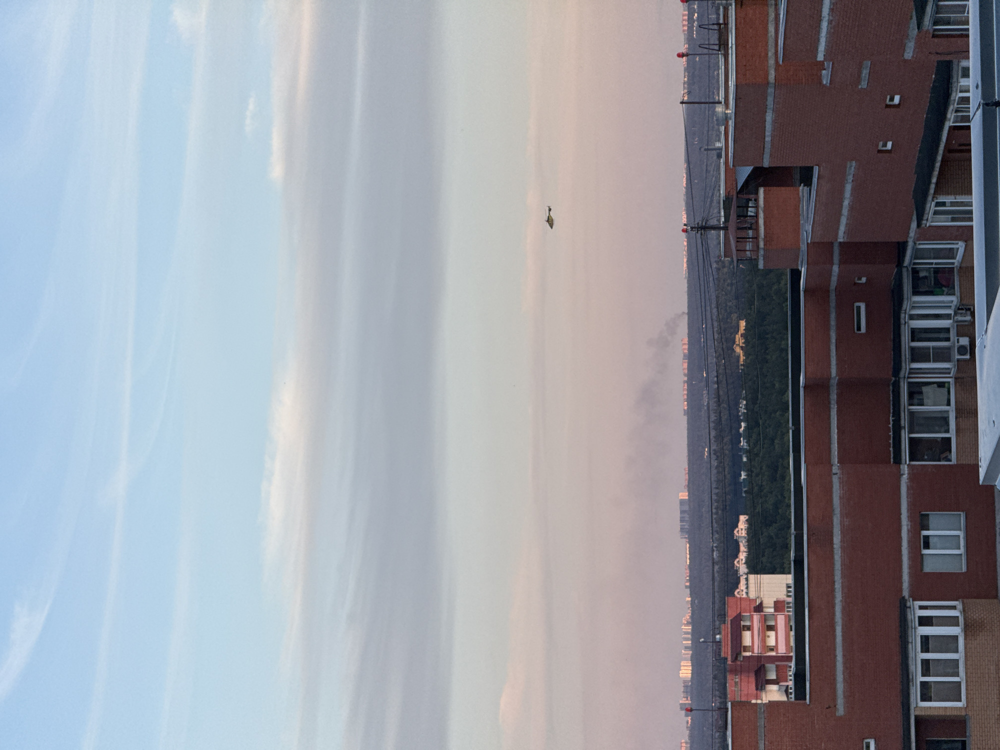 This is a distant aerial view of the city of Khimki, Russia, captured at dusk. The sky is a gradient of pale blue and pink, with wispy clouds. In the mid-distance, a small helicopter is visible flying in the sky. Below the helicopter, the cityscape of Khimki unfolds, characterized by numerous residential buildings and apartment complexes. Some of these buildings are tall, while others are shorter. A patch of dark green forest separates the closer residential areas from the more distant buildings. In the foreground, the rooftops and upper floors of brick apartment buildings are prominent. The scene is observed from a high vantage point, likely from another building, looking out over the urban landscape. There are no visible people or specific actions occurring in the image.