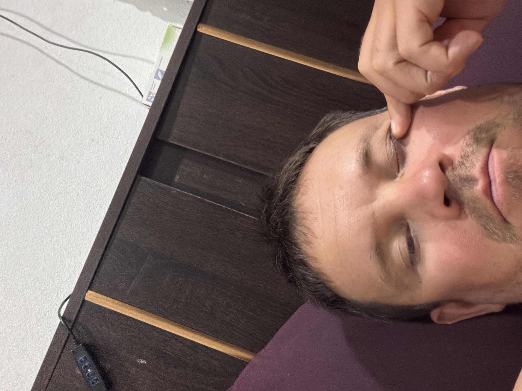 The image shows a close-up of a man's face, with his right eye being pulled open by his finger. He is lying down, with a purple pillow visible to his left. In the background, a dark wooden headboard of a bed is visible. To the top left of the headboard, a small black electronic device with illuminated buttons is attached, and a black cable runs from it towards the right. A white box with some printing on it is also visible on top of the headboard. The man appears to be in a bedroom. No specific details about the location in Baan Mae Nam, Thailand are discernable from the image.