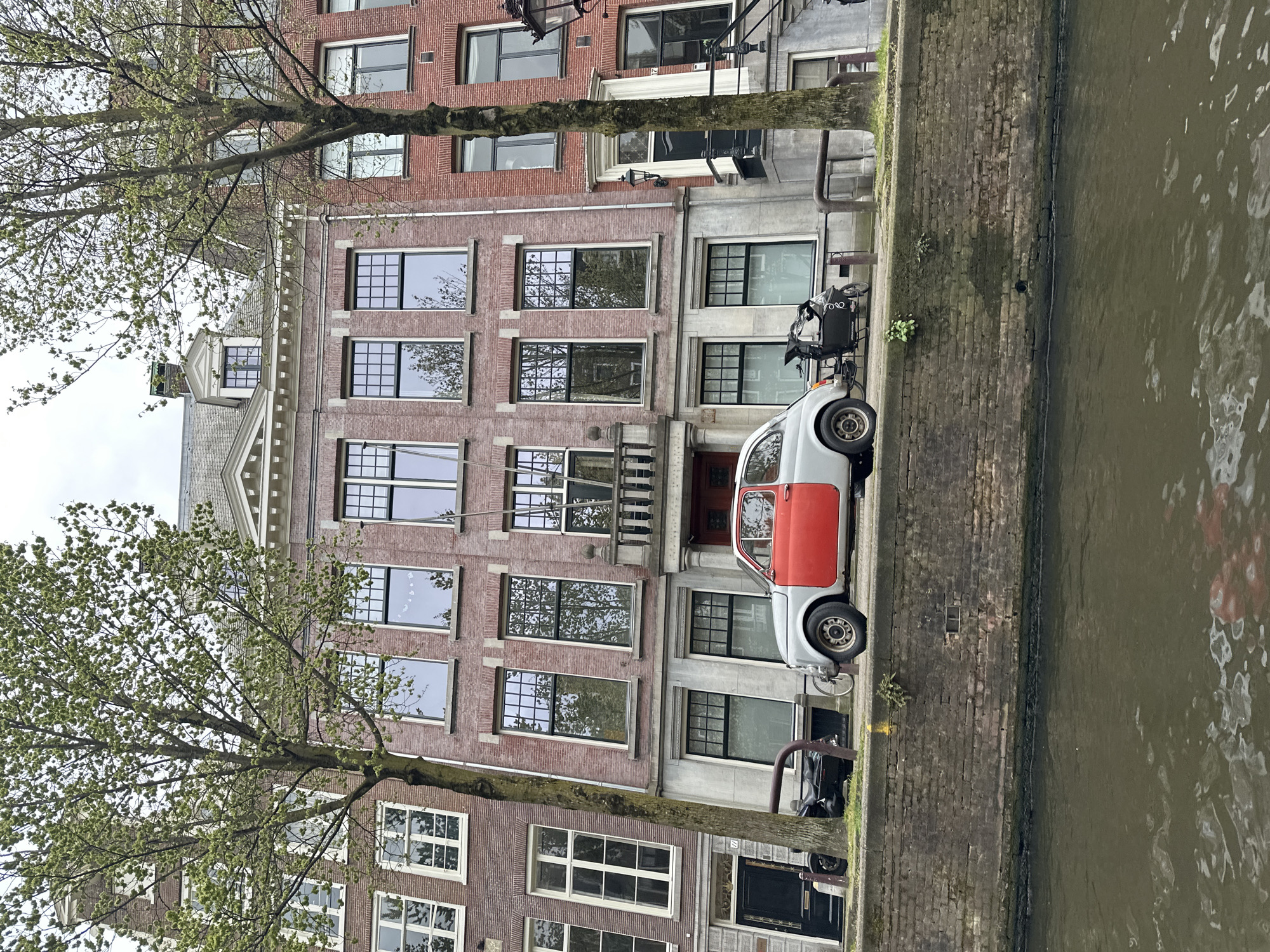 The image depicts a serene canal scene in Amsterdam, Netherlands, under an overcast sky. The view is from the canal, looking towards a row of traditional Dutch canal houses and the adjacent street.
**Subjects and Scene:**
The primary subjects are a series of tall, narrow canal houses, two deciduous trees, a vintage car, and the canal. The setting is a quiet urban street alongside a canal, showcasing typical Amsterdam architecture and atmosphere.
**Buildings:**
Several multi-story canal houses, constructed from reddish-brown brick, line the street. They feature numerous windows, many with white frames and grid patterns, reflecting the sky and surrounding trees. Architectural details include white window sills, decorative cornices, and a prominent stepped gable on the building to the left, adding to the distinctive Amsterdam aesthetic.
**Vehicles:**
A small, light grey vintage car with a contrasting orange-red driver's side door is prominently parked on the street. It resembles a classic Fiat 500. Beside it, a black cargo bike (bakfiets) is also parked, with the letters "EO" visible on its cargo box. Other bicycles and a metal bike rack are partially visible.
**Nature and Canal:**
Two large trees with fresh, light green spring foliage are situated along the street, their branches extending across the upper portion of the image. The canal water is a murky green-brown, displaying ripples and reflections of the buildings and sky. The canal bank is a weathered brick wall, with patches of grass and small plants growing along its surface.
**Notable Details:**
The overcast sky indicates a cloudy daytime. The new, light green leaves on the trees strongly suggest either spring or early summer. A house number "12" is faintly visible above one of the building's entrances. No people are visible in the scene, contributing to a tranquil, everyday street snapshot of Amsterdam.