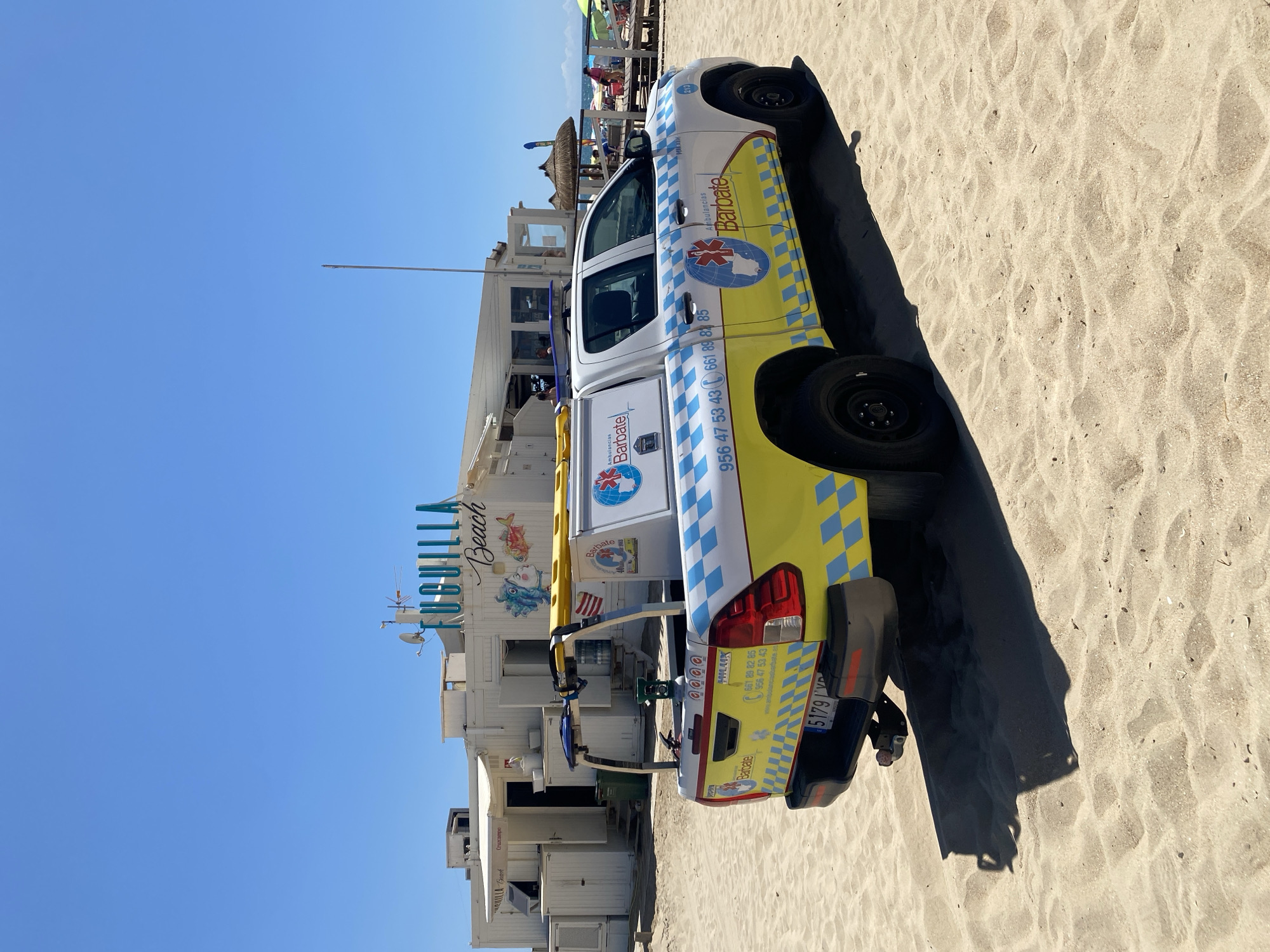 A Toyota Hilux pickup truck, marked with "Ambulancias Barbate," is parked on the sandy beach of Fuguilla Beach, in the city of Conil de la Frontera, Spain. The truck is white with yellow and blue checkered accents, and features medical symbols and phone numbers. In the background, a white building with "Fuguilla Beach" signage is visible, along with beach umbrellas, chairs, and a few people relaxing under them. The sky is clear and blue, and the overall scene depicts a sunny day at the beach.