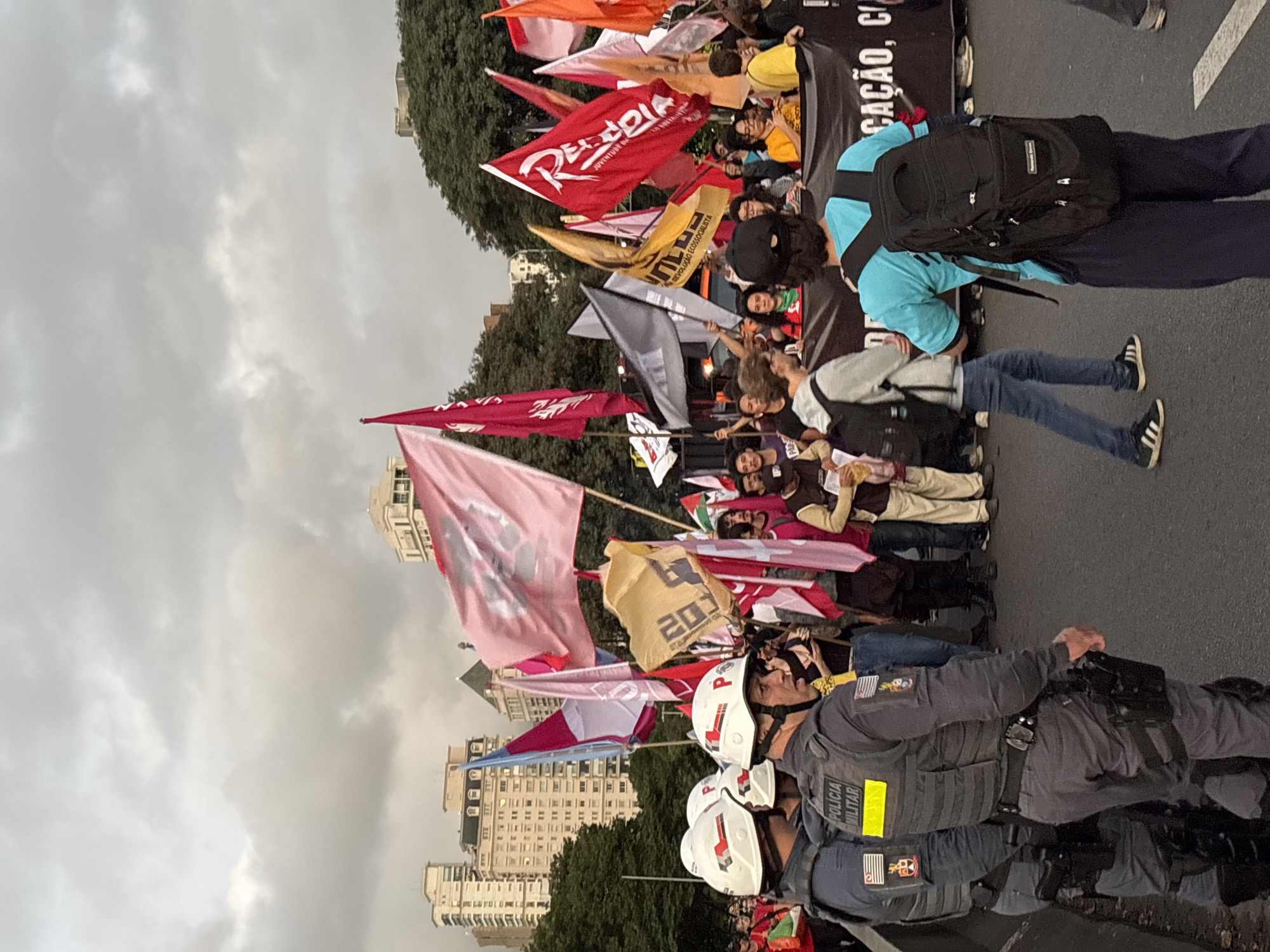 The image depicts a street scene with a protest or demonstration in progress. The foreground features several police officers, clad in dark gray uniforms and white helmets, facing toward the crowd. The officers are positioned in front of a large group of people holding various flags and banners.
The flags display different colors and symbols, some with text in Portuguese. One flag is red with the word "Rede Pela Democracia Juventude Socialista" written on it, another says "MULEE Revolução Ecosocialista". A large black banner is visible behind the crowd. The crowd contains a diverse mix of people.
In the background, a series of tall buildings with symmetrical windows are visible, indicating an urban environment. The sky is overcast with gray clouds.
Based on the attire of the police officers, specifically the patch on their uniforms, this scene is most likely taking place in the city of São Paulo, Brazil.
