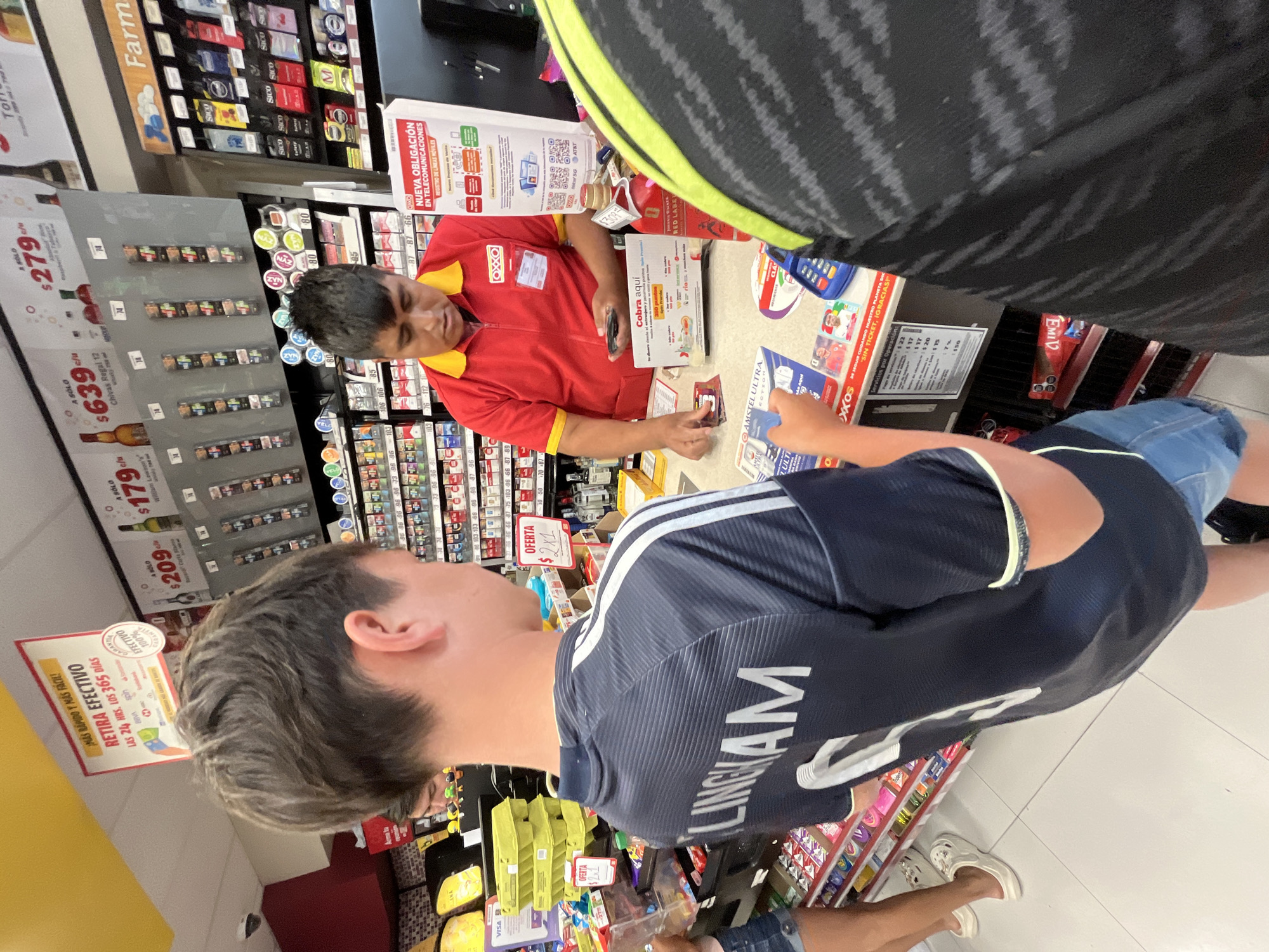 The image shows a customer paying at a cash register inside an OXXO convenience store. The customer, a young man wearing a navy blue soccer jersey with "LINGHAM" printed on the back and denim shorts, is holding payment in his hand for the cashier, a man in a red OXXO uniform. The cashier is taking the payment. Various products are visible on the shelves behind the cashier, including cigarettes, cigars, alcohol, and condoms. Prices are displayed on shelf tags. A yellow sign advertises cash withdrawals. The store appears to be in San Bernardino Tlaxcalancingo, Mexico, as evidenced by the OXXO branding and product labeling in Spanish.