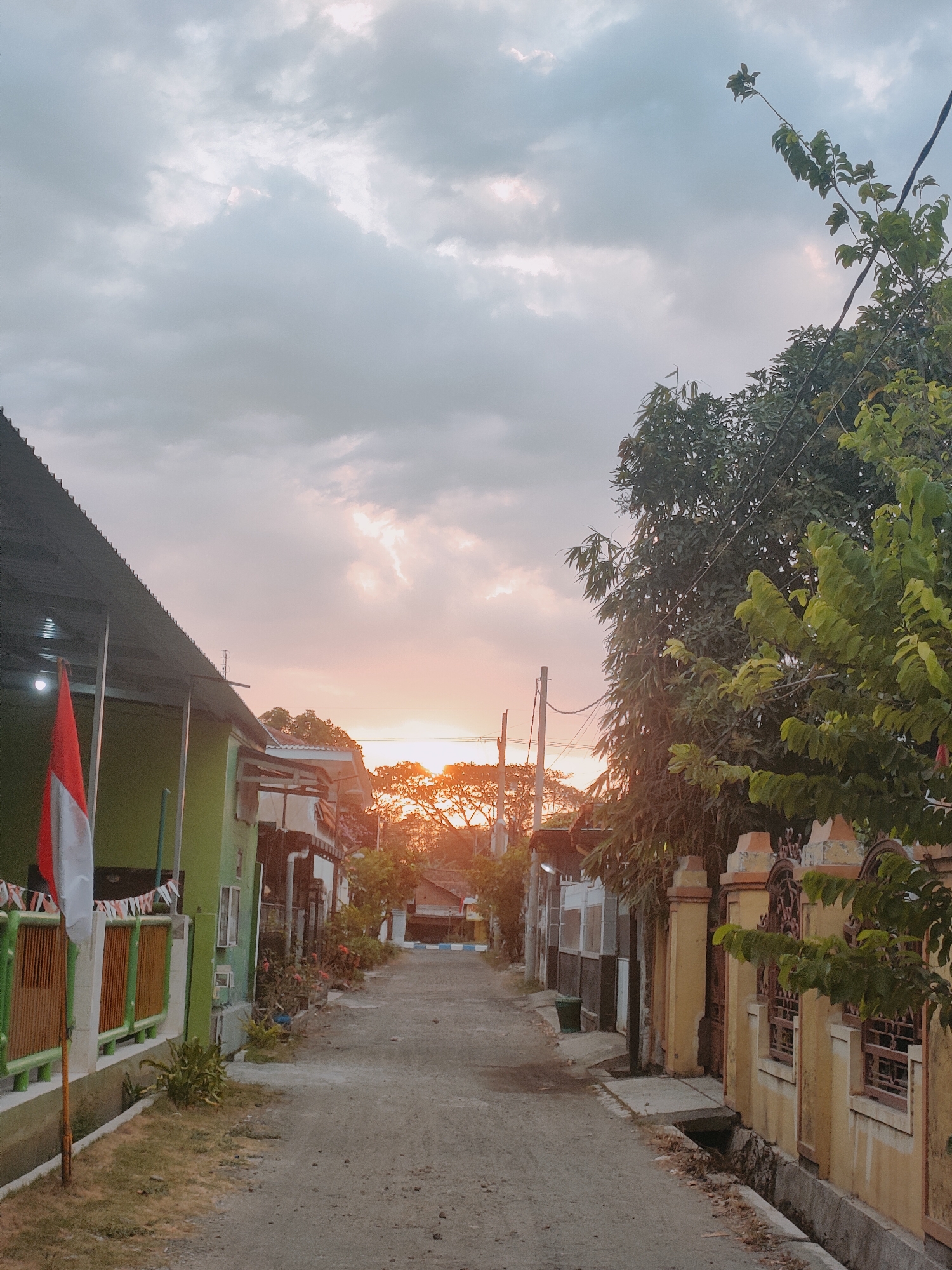 The image depicts a residential street in Sooko, Indonesia, during a sunset. The sky is filled with dramatic clouds, with the warm glow of the setting sun visible between them. On the left side of the street, a two-story green house is partially visible, with an Indonesian flag prominently displayed in front. The house has a covered porch and a fence. Further down the street, other houses and buildings can be seen, lining both sides of the road. The street itself is paved with gravel and dirt. Lush green trees and foliage are present on the right side of the street, framing the view and casting shadows. The overall atmosphere is serene and peaceful, capturing the quiet end of the day in a suburban setting.