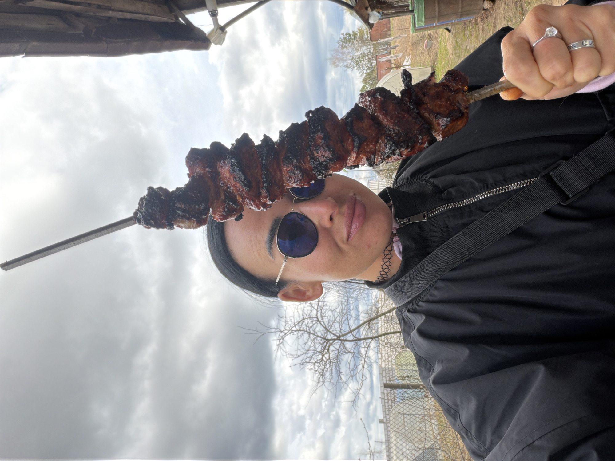 A person with sunglasses and a black jacket holds a skewer of grilled meat. The person is wearing rings on their fingers and a choker necklace. The background includes a cloudy sky, trees, and a fence. The location appears to be outdoors, in the city of Бронница, Russia.