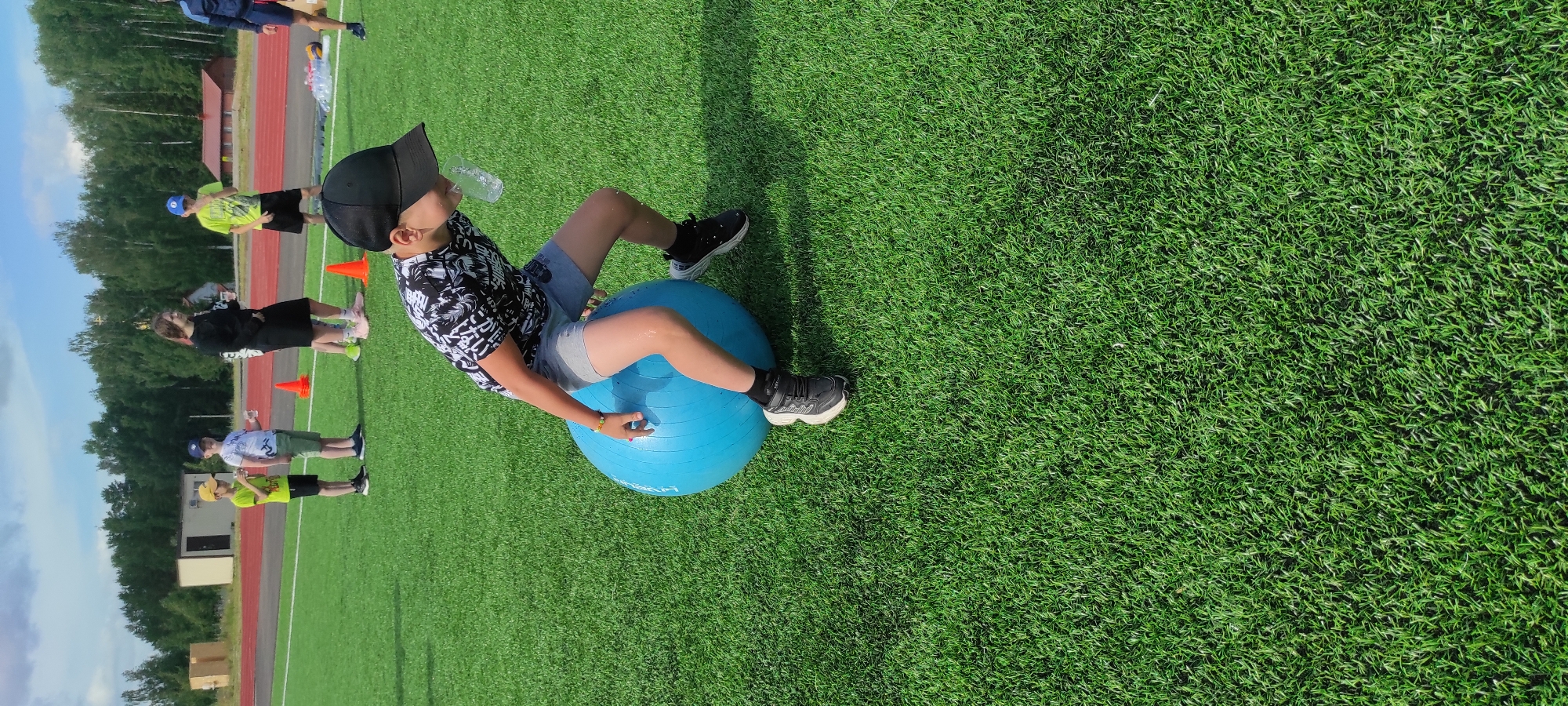 In Noviy, Russia, on a bright, overcast day, a young boy with a black baseball cap and a black and white patterned t-shirt is participating in an outdoor activity on a vibrant green artificial turf field. He is perched precariously on a large, blue exercise ball, leaning forward as if engaged in a game of balance. A clear plastic cup is held to his lips, suggesting he might be attempting to drink or carry liquid without spilling. His grey shorts and black athletic shoes complete his outfit.
In the background, other children and adults are visible. Two young boys, one in a yellow t-shirt and the other in a white t-shirt, stand with drinks in hand, possibly waiting for their turn or observing the activity. A young woman in a black dress and a man in a blue athletic jacket are also present, along with other figures further in the distance. Orange traffic cones are scattered on the field, indicating a designated course or area for the event. Behind the field, a treeline forms a natural backdrop, and a few buildings, including a small, light-colored structure and a more distant red building, are visible. The scene suggests a casual, organized event, possibly a school or community gathering, taking place at an outdoor sports facility.