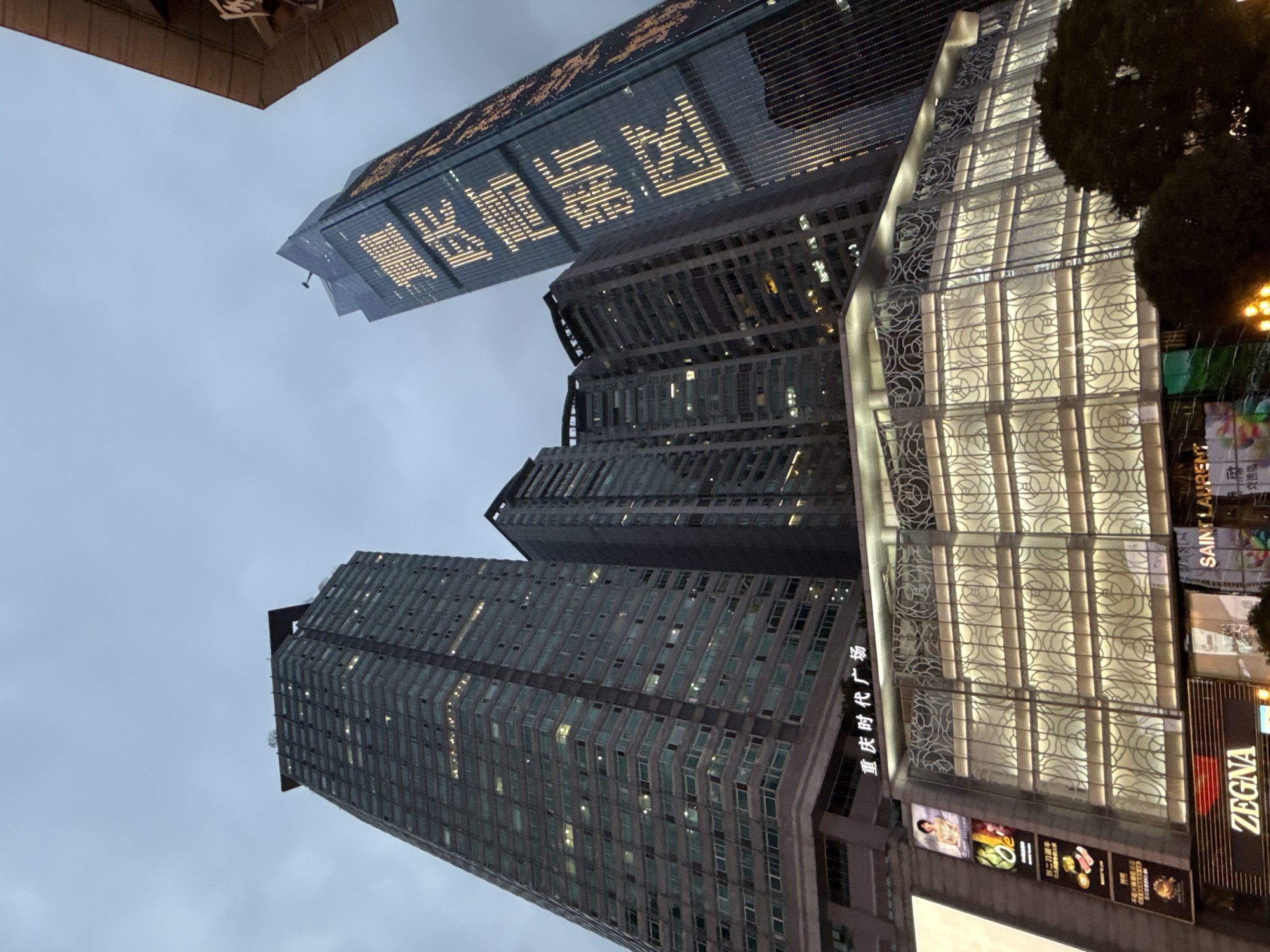 This image depicts a nighttime urban scene in Yuzhong District, China, focused on a cluster of modern skyscrapers and a multi-story building with illuminated facades. The sky is overcast and dark, suggesting either dusk or night.
The most prominent structure is a tall, dark skyscraper on the right, its glass facade illuminated with large, bright yellow characters that appear to be Chinese script. Below this, a lower, glass-fronted building is lit from within, displaying decorative patterns on its exterior. Signs for "SAINT LAURENT" and "ZEGNA" are visible at the base of this structure, indicating a retail or commercial area.
To the left, another large skyscraper is visible, its exterior also featuring numerous illuminated windows, suggesting occupied floors. A sign at the base of this structure reads "重庆时代广场" (Chongqing Times Square) in Chinese characters. Several smaller buildings and a portion of another skyscraper are also present in the background.
The overall impression is one of a bustling, contemporary urban environment with a strong emphasis on commercial activity and modern architecture. The lighting from the buildings creates a dramatic contrast against the dark sky. No people are clearly discernible in the image.