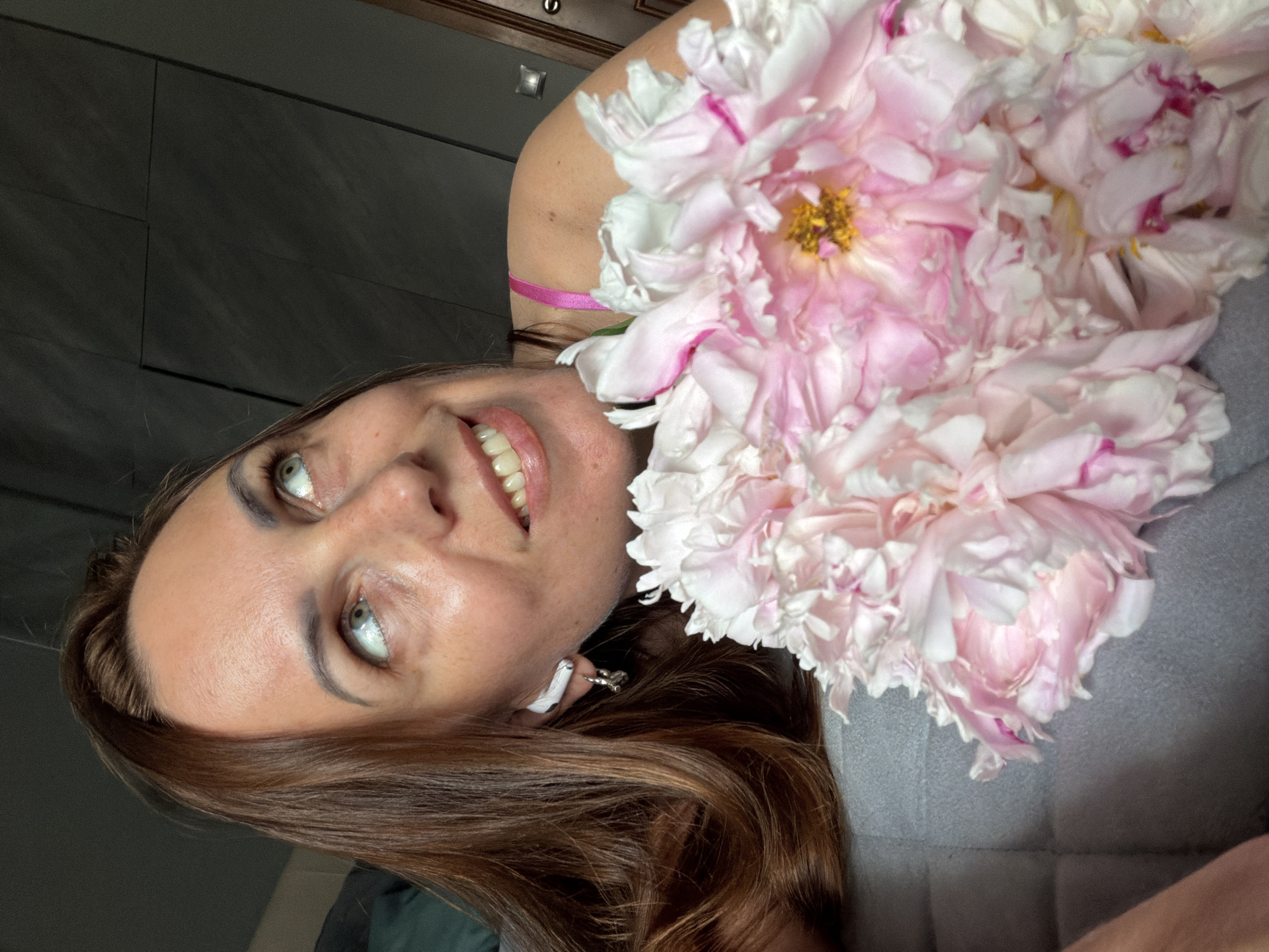 In the foreground, a bouquet of pink and white peonies rests on a gray, quilted surface. A woman with long, brown hair is partially visible, lying down and holding the flowers. Her head is tilted upwards and to the right, with her eyes looking in the same direction. She is wearing white earbuds in her left ear. Her skin appears smooth, and she has a light makeup application. Her mouth is slightly open in a smile. The background is dark and indistinct, suggesting an indoor setting. There is no indication of the city of Birobidzhan, Russia.