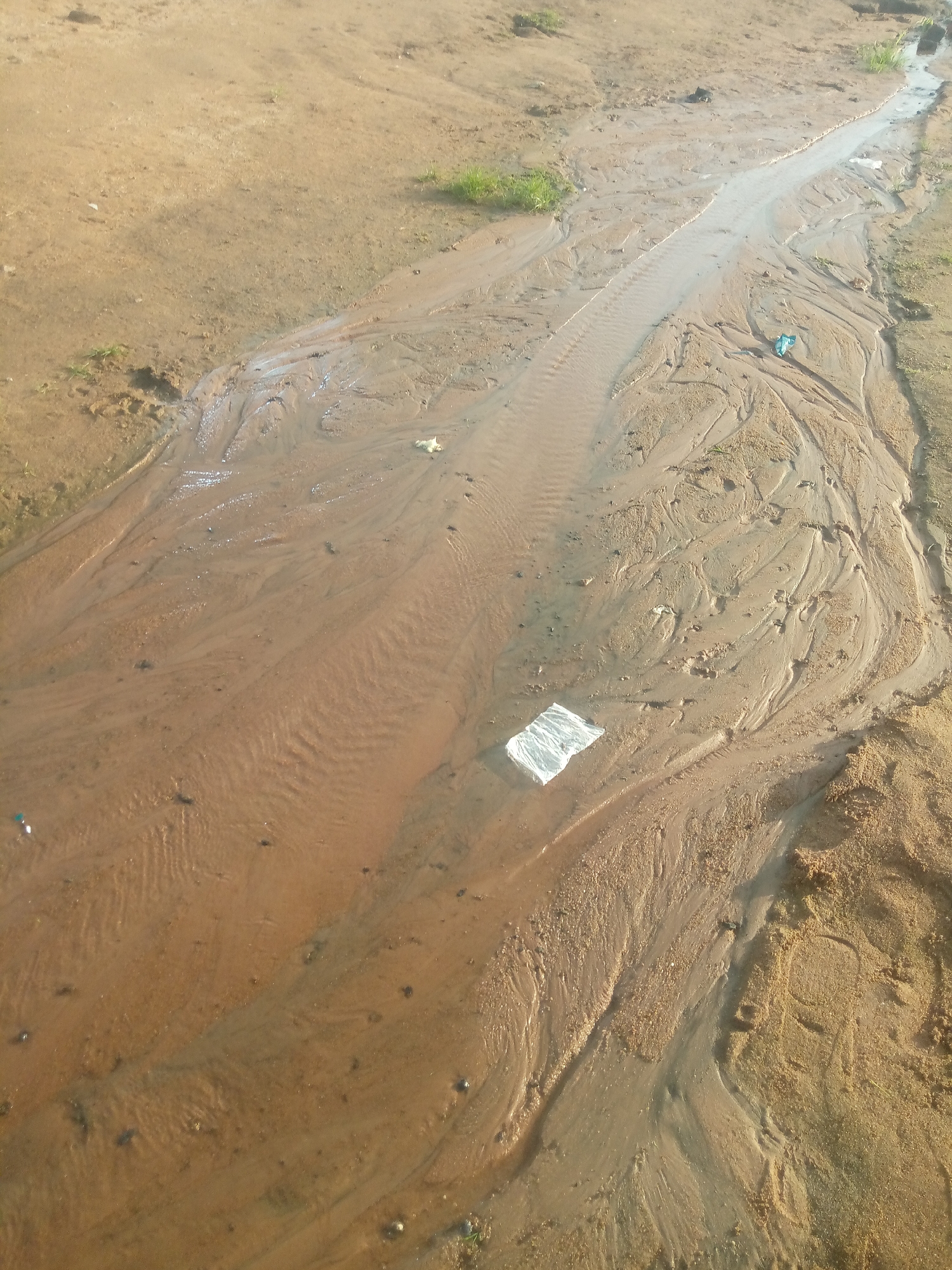 The image depicts a shallow stream of water flowing over sandy ground, creating natural channels and patterns in the soil. A piece of white plastic debris is visible in the stream. The environment appears to be a natural area, possibly a riverbed or a drainage channel, with sparse vegetation on the higher ground. The lighting suggests it is daytime.
