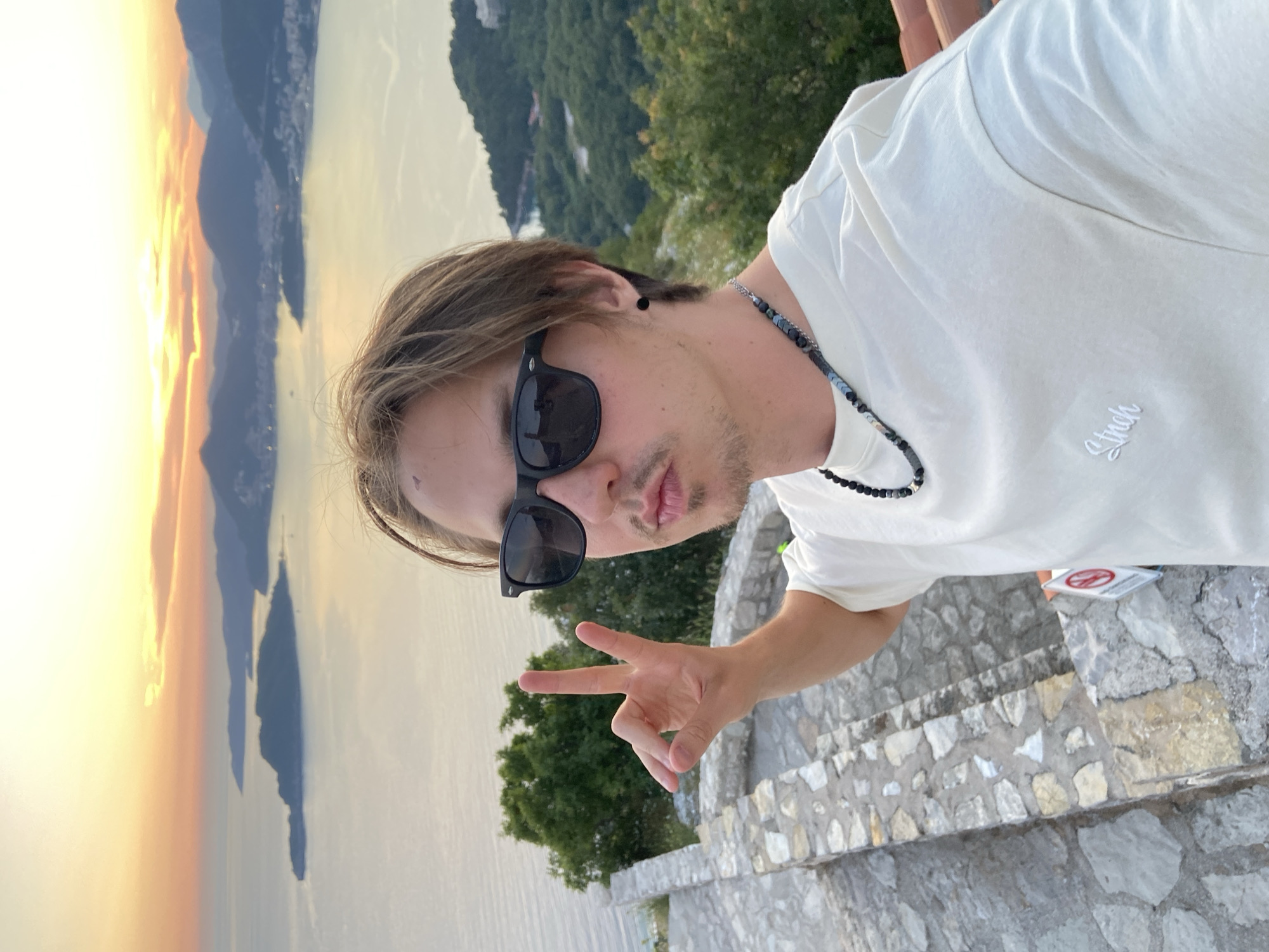 A young man is posing for a selfie outdoors, likely on a stone staircase or ledge, given the visible stone construction. The subject is wearing a white t-shirt, sunglasses, and a beaded necklace. He is making a peace sign with his left hand and has a neutral expression. The background reveals a scenic coastal view, with water, distant land formations, and a sunset. The location is identified to be in Sveti Stefan, Montenegro based on the coastline and the appearance of the landscape. The angle and composition suggest the photo was taken with a handheld device.