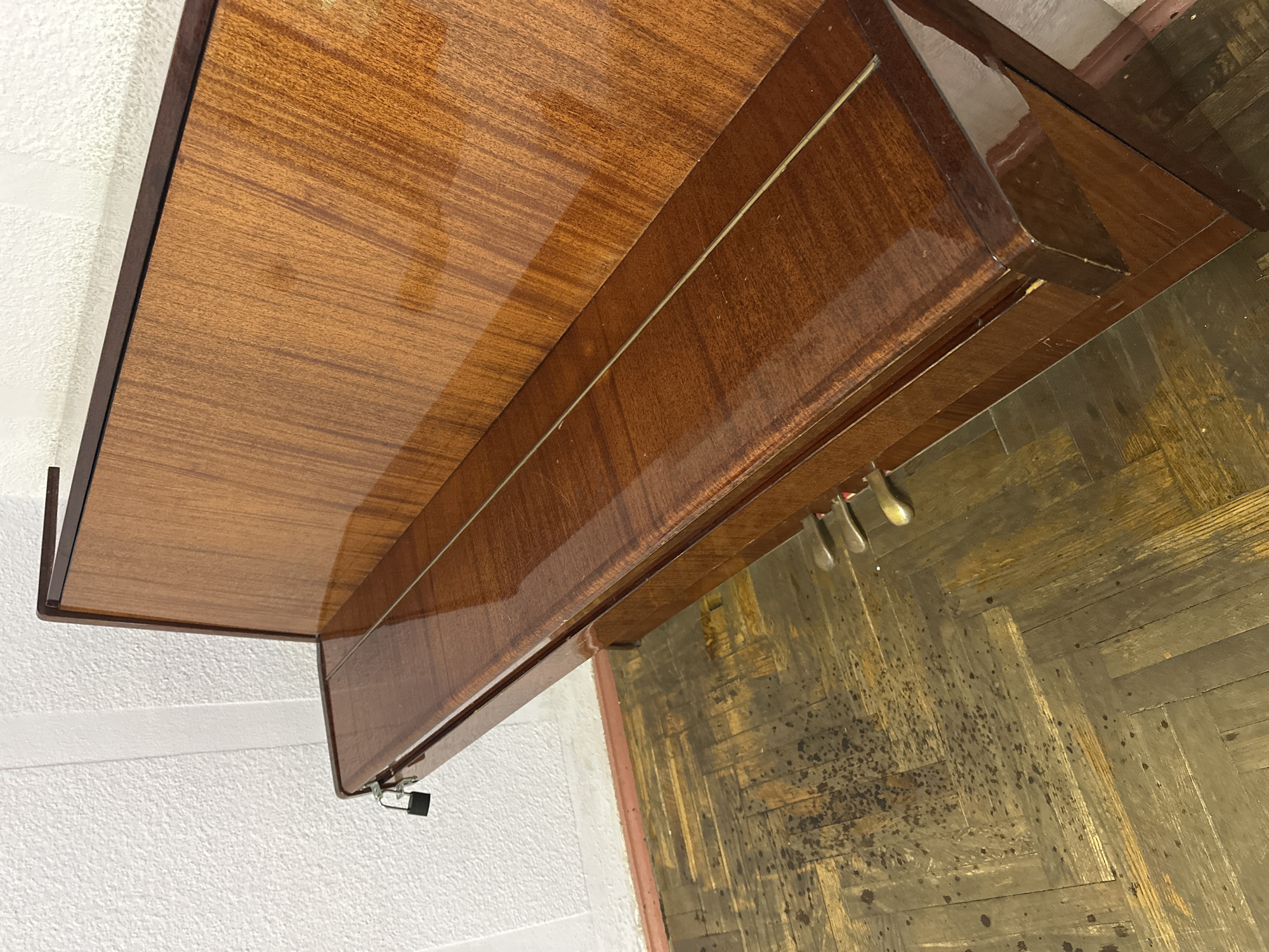 The image shows the keyboard and pedals of an upright piano. The piano has a polished wooden finish and is situated indoors, against a white textured wall. A small black padlock is attached to the edge of the piano. The floor is a parquet pattern with visible wear and discoloration. The scene is devoid of any people. The location is stated to be in Ракаўскі сельскі Савет, Belarus.