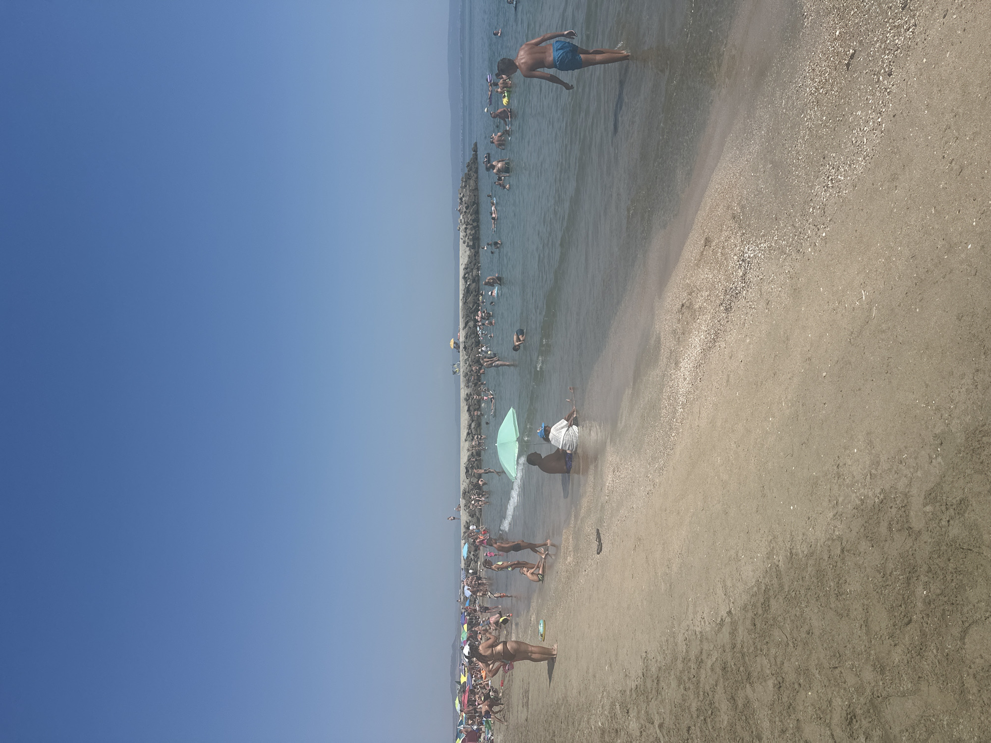 This image shows a crowded beach scene on a sunny day in Pomorie, Bulgaria. The foreground is dominated by a sandy beach with some shells and footprints. In the middle ground, people are wading in the shallow water, swimming, and relaxing under colorful umbrellas. A stone breakwater extends into the sea in the background, where more people are gathered and in the water. The sky is clear and blue. The overall scene depicts a typical summer day at a popular tourist destination.