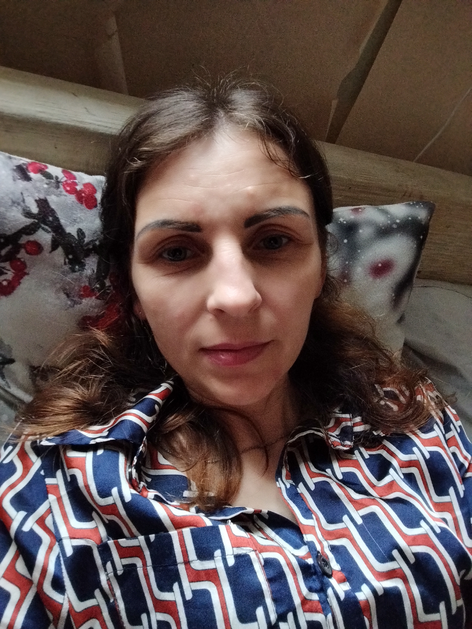 A medium shot, indoor, eye-level selfie of a woman lying down, resting her head on pillows. The woman is facing the camera, with her dark brown hair spread around her head. She is wearing a blue, white, and red geometric patterned blouse. Her expression is neutral. The background consists of wooden paneling and two pillows, one with a floral pattern and the other with a dark pattern with red and white elements. There is no specific indication of location in Vinnytsia, Ukraine.