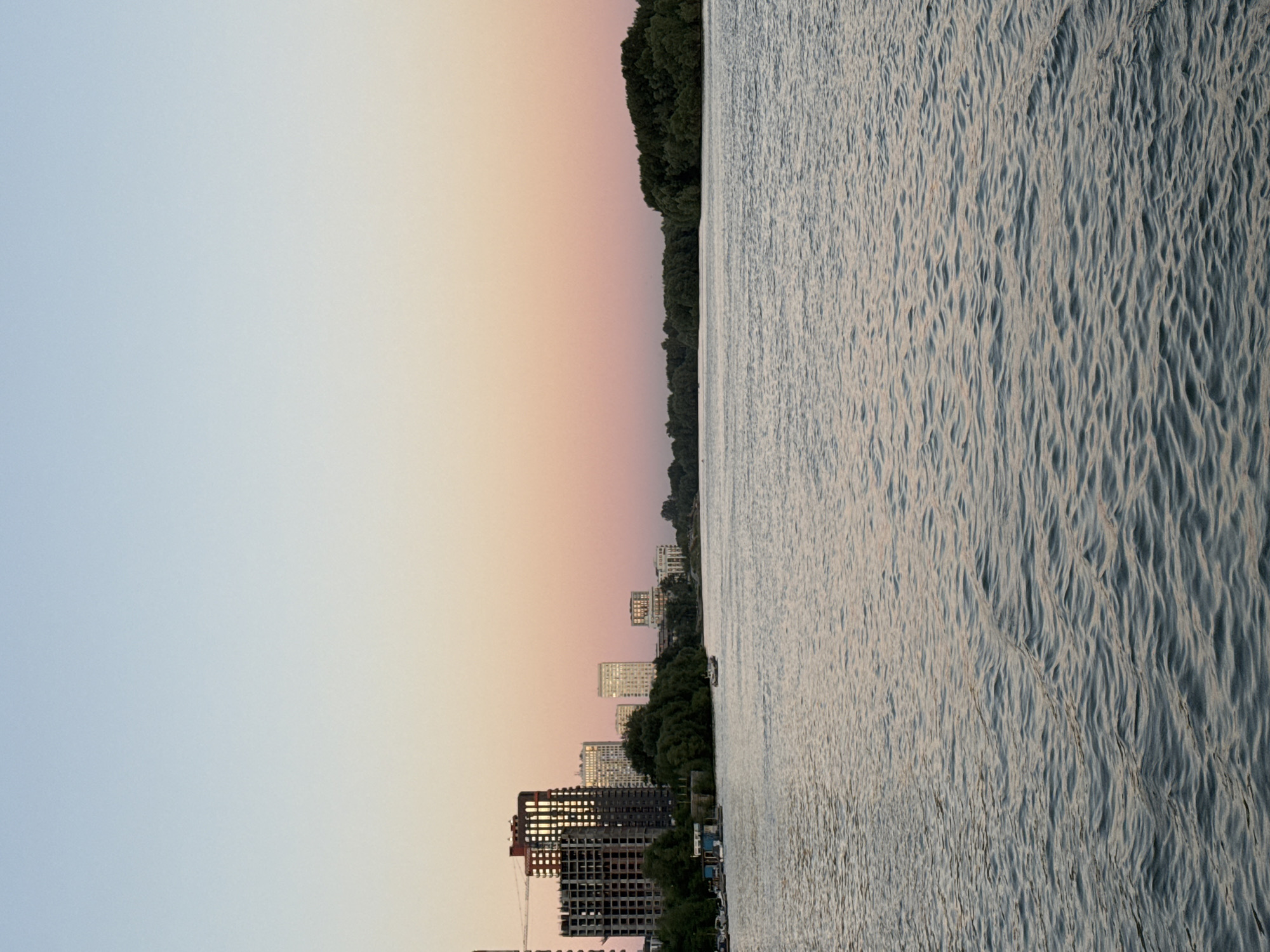 The image captures a serene sunset over the Ob River in Novosibirsk, Russia. The sky transitions from a pale blue at the top to a soft pink and orange hue near the horizon. On the left bank of the river, a cluster of modern buildings, some under construction and others completed, are visible, interspersed with green trees. A few boats are moored along the shore. The river itself occupies the foreground, its surface rippled by a gentle breeze, reflecting the soft colors of the sky. On the right side of the river, a dense line of trees creates a natural embankment. The overall scene is tranquil, showcasing an urban landscape harmoniously integrated with a natural waterway during the twilight hours.