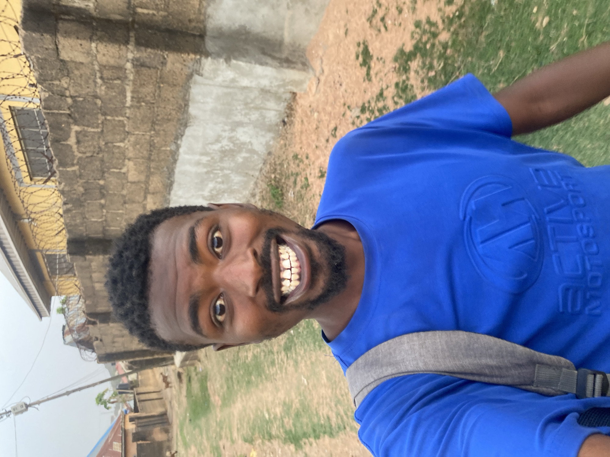 An upright, eye-level selfie captures a young man outdoors in Barakin Akawo, Nigeria, smiling broadly at the camera. He has dark skin, a full beard, and short, dark curly hair. His eyes are wide and bright, and his cheerful, open-mouthed smile reveals a full set of teeth.
The man is wearing a bright blue, short-sleeved t-shirt. A grey backpack strap is visible across his chest and left shoulder. The t-shirt features embossed text on the front, which clearly reads "ACTIVE MOTOSPORT".
The background suggests a semi-urban or residential area under diffused daylight, possibly an overcast or hazy sky. To the man's left, a wall constructed from dark, irregular stone or concrete blocks is visible. Above this wall, a lighter-colored building features barbed wire along its upper edge and a small, barred window. To his right, the ground transitions from a patch of dirt with sparse grass to a more barren, light brown dirt area. In the distance, various buildings, a utility pole with overhead wires, and scattered vegetation can be seen, indicating a populated environment. The overall atmosphere conveyed by the man's expression is one of happiness or contentment.