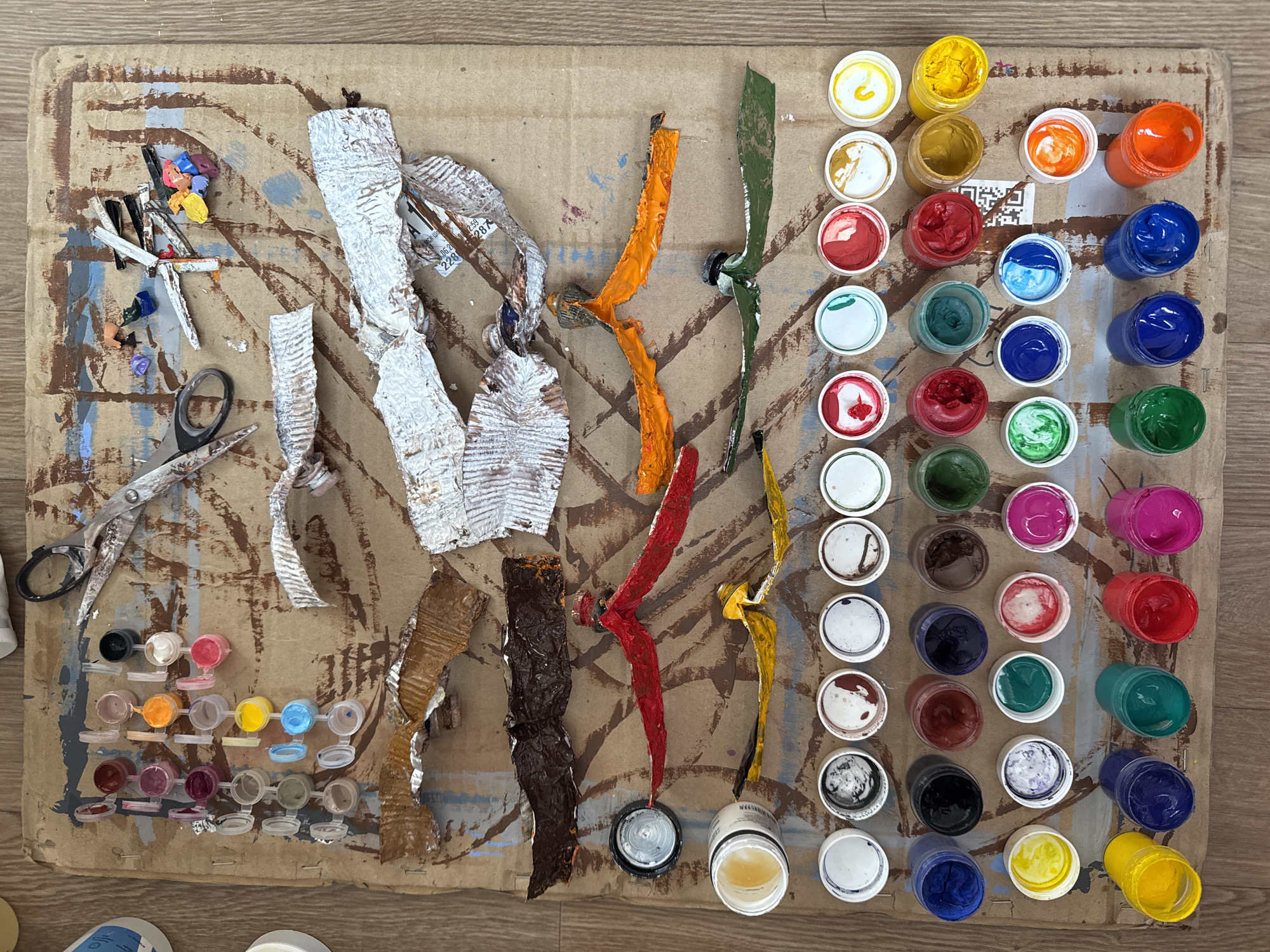 A high-angle shot shows a paint-covered piece of cardboard serving as a makeshift art workspace. The cardboard exhibits a chaotic arrangement of various items. On the top left is a collection of painting tools and a pair of scissors. Across the center of the board, there is a grouping of flattened, used paint tubes in assorted colors, including silver, brown, red, orange, green, and yellow. To the right, there are numerous small, open containers of paint in an array of colors, including yellow, orange, red, blue, green, brown, white, black, and pink. The image does not contain any people. There are no identifiers to determine the location of Moscow, Russia.