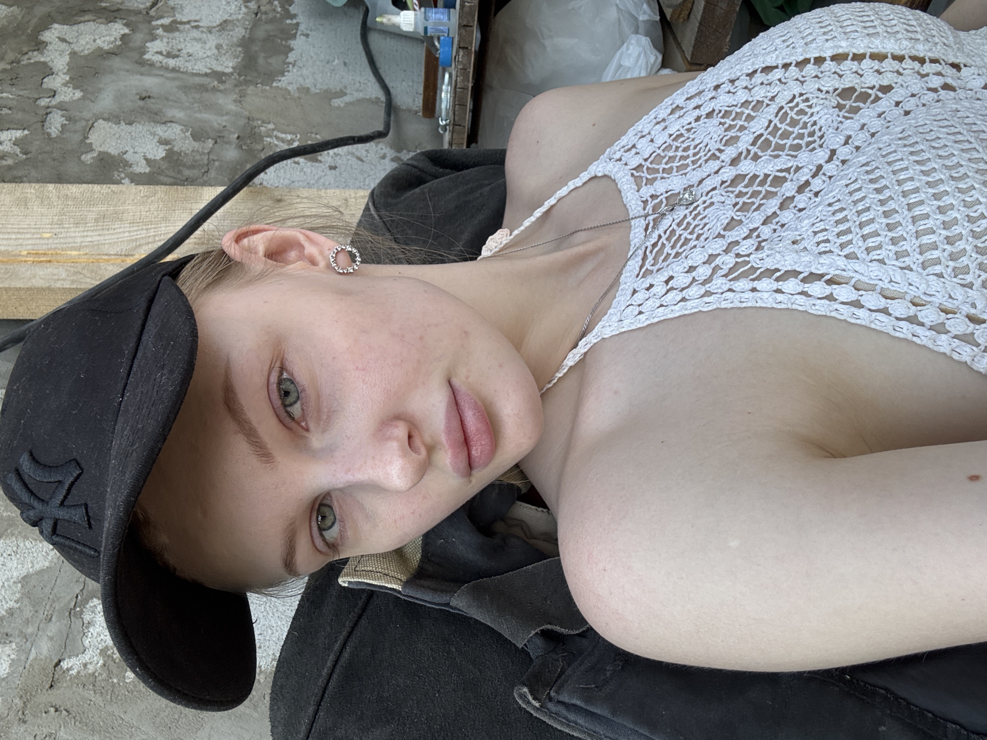 A young woman is lying down, viewed from an angle. She is wearing a black baseball cap with the "NY" logo, a white crocheted top, and a necklace. She has fair skin, freckles on her face, and green eyes. The background consists of a concrete wall, a wooden plank, and some equipment. The image appears to be taken outdoors during the day, but it is difficult to determine the exact location in Perm, Russia without more context.