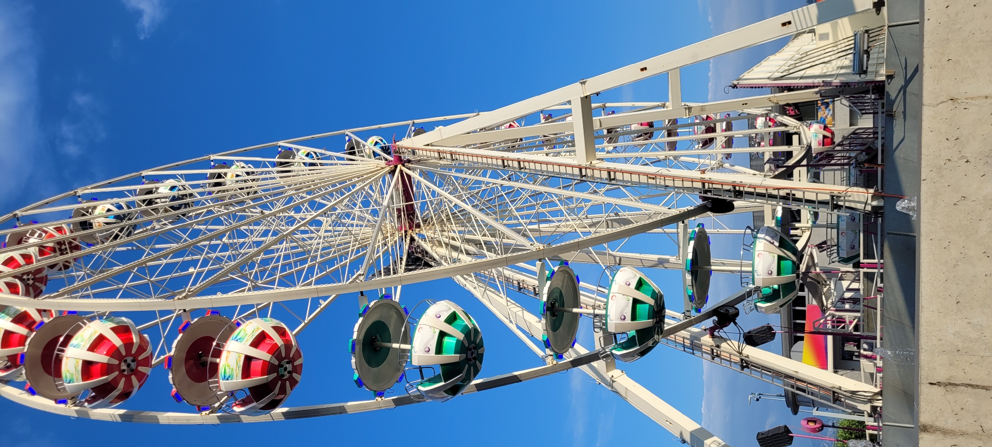 A large white Ferris wheel stands tall against a clear blue sky with a few wispy clouds. The Ferris wheel has numerous gondolas, some of which are visible from below, with red and white patterns and green and white patterns. The gondolas are illuminated with blue lights. A metal ladder runs vertically up the side of the Ferris wheel structure. In the foreground, a concrete barrier runs horizontally across the bottom of the image, with small fountains of water splashing from it. Behind the barrier, there is a section of the amusement park with some structures and colorful lighting. The Ferris wheel appears to be part of a fairground or amusement park located in Rapperswil-Jona, Switzerland, given its typical use in such locations.