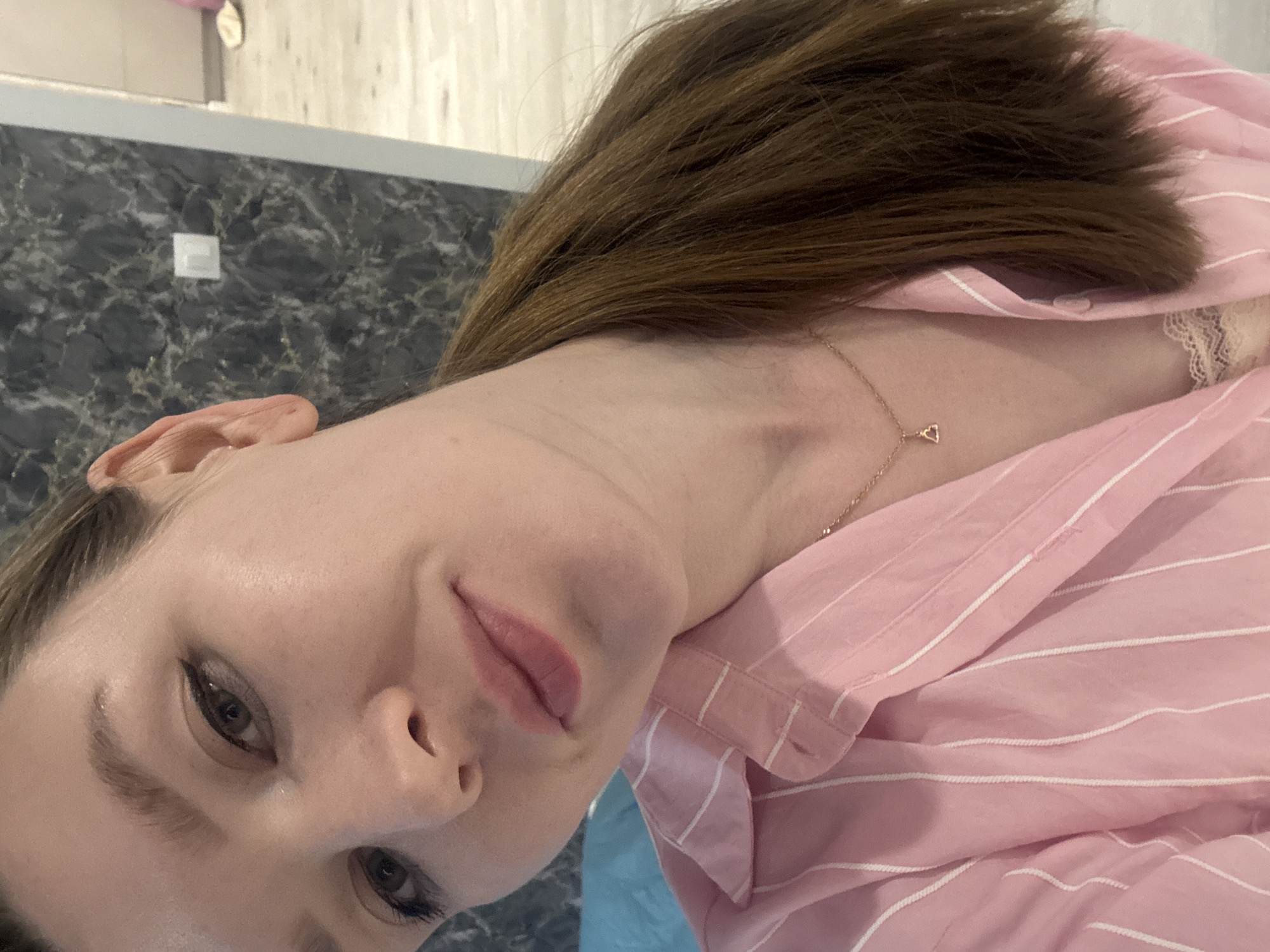 The image shows a close-up of a woman's face and upper torso. She has fair skin, brown hair, and is wearing a pink and white striped shirt. A delicate gold necklace with a small pendant is visible around her neck. Her expression is a slight smile. The background includes a textured wall, possibly marble, with a light switch visible on it. Part of a blue object is also in the lower background. The location is not determinable from the image, and therefore it's impossible to assert it is in Izhevsk, Russia.