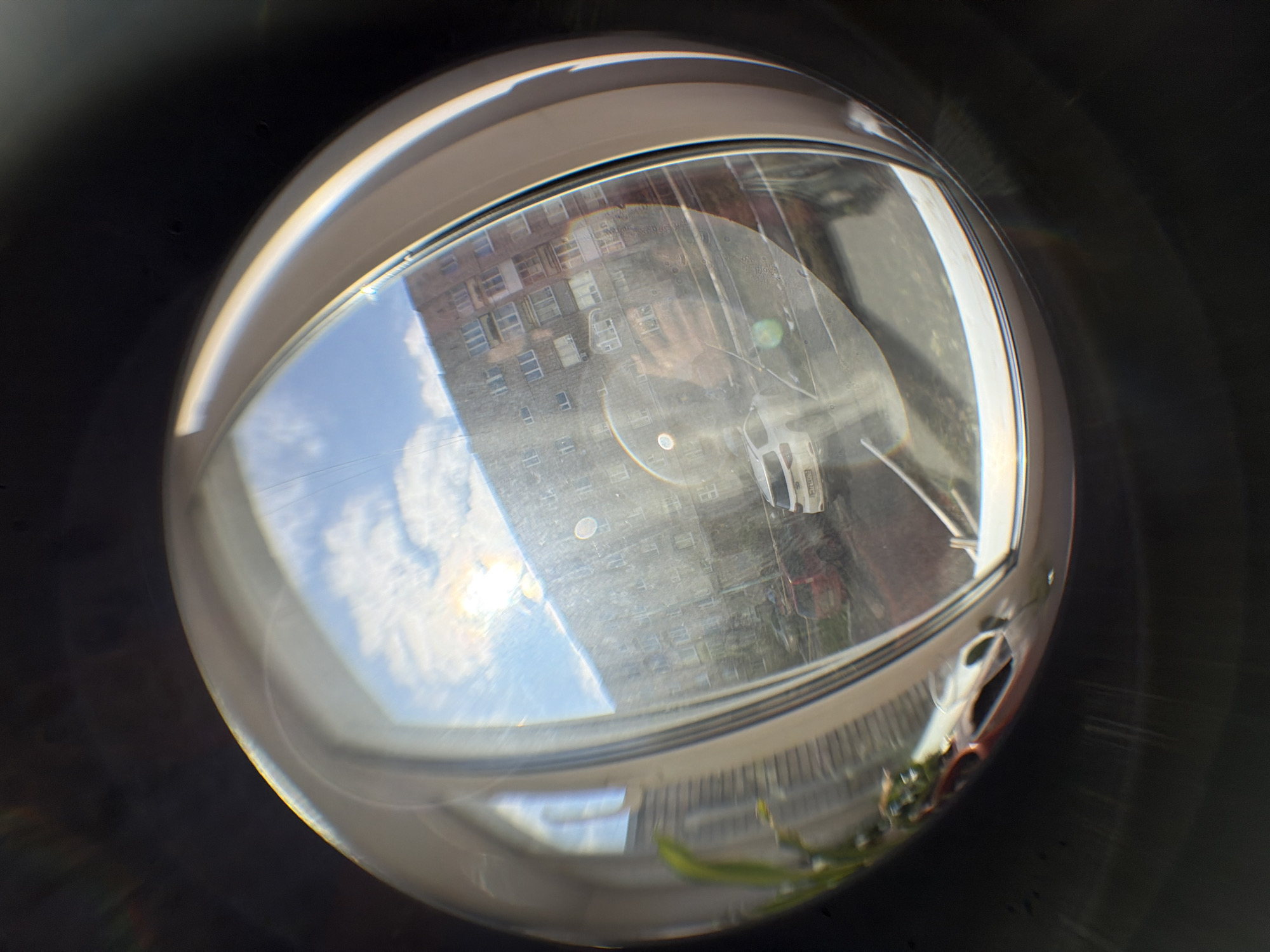 The image appears to be a fisheye lens view of a cityscape reflected in a glass orb or lens. A tall apartment building with multiple rows of windows dominates the reflection. The sky above the building is visible, showing a mix of blue sky and white clouds. A street with vehicles is also reflected. The orb itself sits on a surface, with some foliage visible along its lower edge. Based on the style of apartment building and the presence of certain vehicles, the location is likely Izhevsk, Russia.