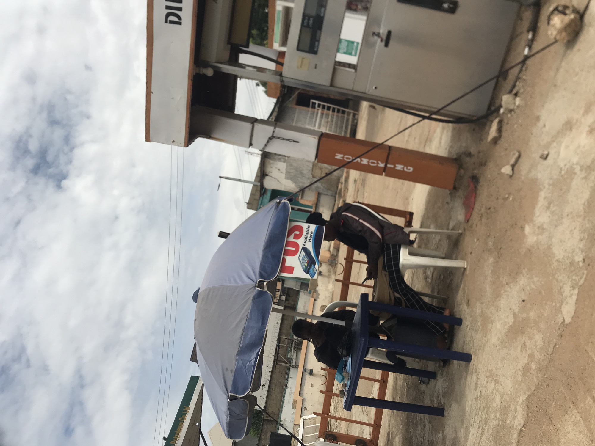 The image depicts an outdoor scene in Rafin Par, Nigeria, under an overcast sky, focusing on two individuals seated beneath a large umbrella near a petrol station.
A dark-skinned individual wearing a dark long-sleeved top is seated on a white plastic chair at a blue plastic table, facing the right side of the frame. Items, including a dark object and light-colored materials, are present on the table in front of them. To their right, a second dark-skinned individual is seated on another white plastic chair, wearing a dark jacket with white stripes on the sleeves and plaid trousers, and actively viewing a mobile phone.
A large, two-toned grey and blue umbrella provides shade over the table and individuals. Behind the individuals and to the right, a red and white sign, partially visible, advertises "POS Available Here" with an image of a POS terminal. To the far right, a silver-grey petrol pump with a digital display is visible beneath a white canopy structure with an orange trim, displaying the partial text "DI" (likely "DIESEL"). Adjacent to the pump, an orange vertical pillar clearly displays "NO SMOKING".
The ground is a light brown-grey concrete or paved surface, showing various textures and minor debris. In the background, several buildings of varying construction are visible, along with a low, light-colored barrier behind the table setup. Overhead power lines are visible against the cloudy sky. The overall lighting is soft and diffuse, consistent with an overcast day.