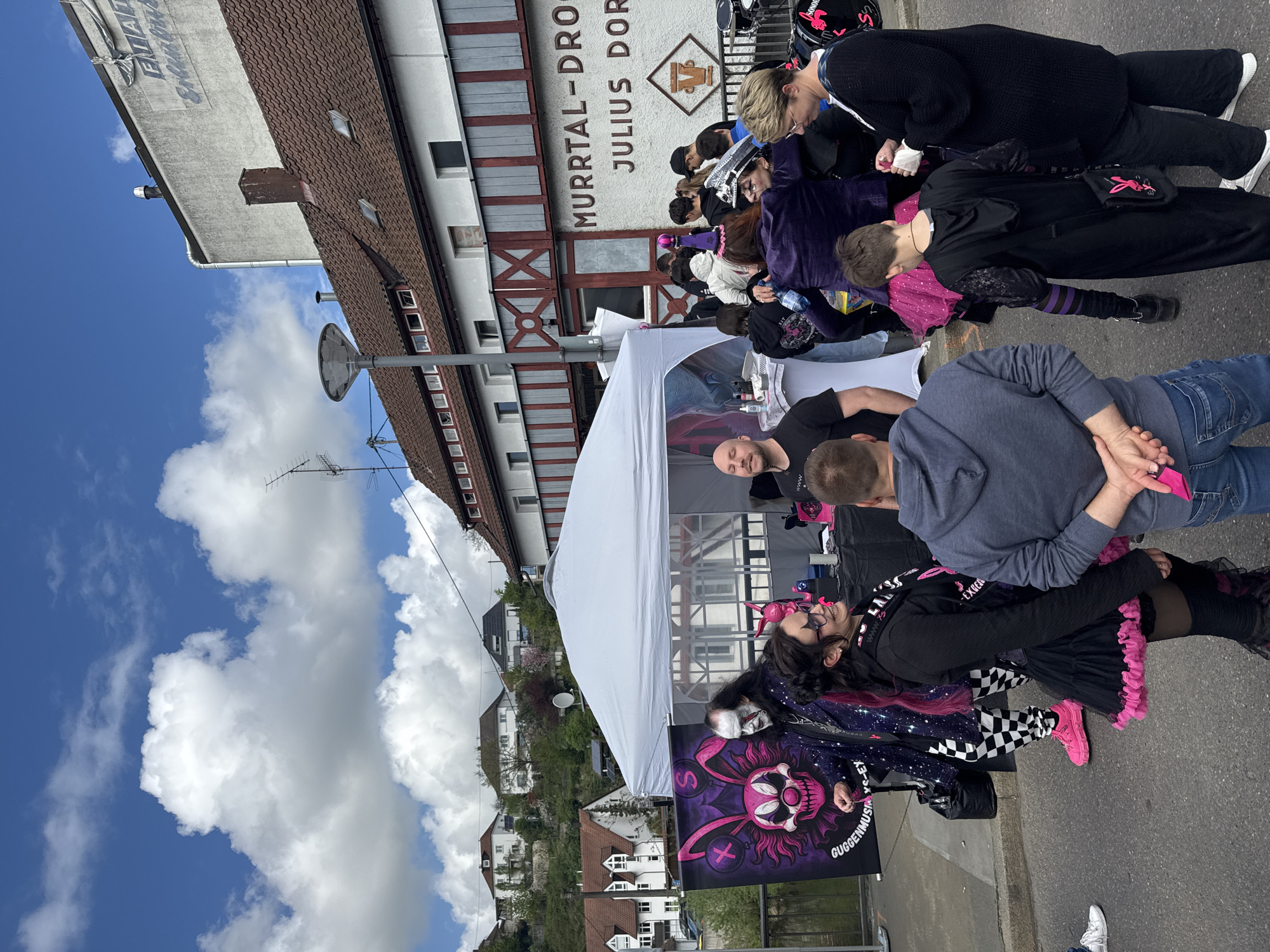 Captured at a slight oblique angle, the image features a gathering under a white tent on a road. Several individuals stand and interact around the tent. In the foreground, a banner with a skull design and the text "GUGGENMUSIXS-EY" is visible. Buildings line the background, with the sign "MURRTAL-DRO JULIUS DOR" prominently displayed on one building. The setting appears to be Backnang, Germany, given the signs in German and architecture. The sky, partially visible, reveals a mix of blue and cloudy weather.