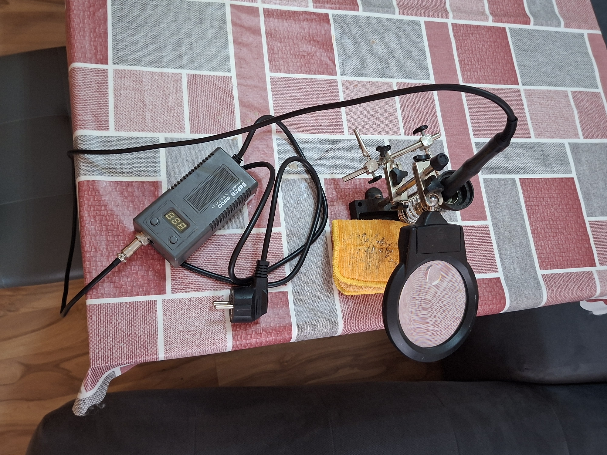 An overhead shot captures a home electronics workbench setup on a patterned tablecloth in what appears to be an indoor setting in Plovdiv, Bulgaria. The central item is a gray BAKON 950D portable soldering station, clearly labeled with its model and function, featuring a digital display currently showing "888". Cables emanate from the station, connecting to a European CEE 7/7 type power plug resting on the tablecloth. To the right, a set of "helping hands" stands ready, equipped with multiple adjustable clamps and an attached circular magnifying glass, suggesting close-up work. A small, crumpled orange cloth, possibly for cleaning soldering tips, is placed beneath the helping hands. The tablecloth displays a repetitive pattern of red, gray, and white squares. Portions of a dark, upholstered sofa or chair are visible along the right and bottom edges, while a wooden floor peeks out in the lower left corner. The scene is static, depicting the tools arranged for use, but no active work or person is present.