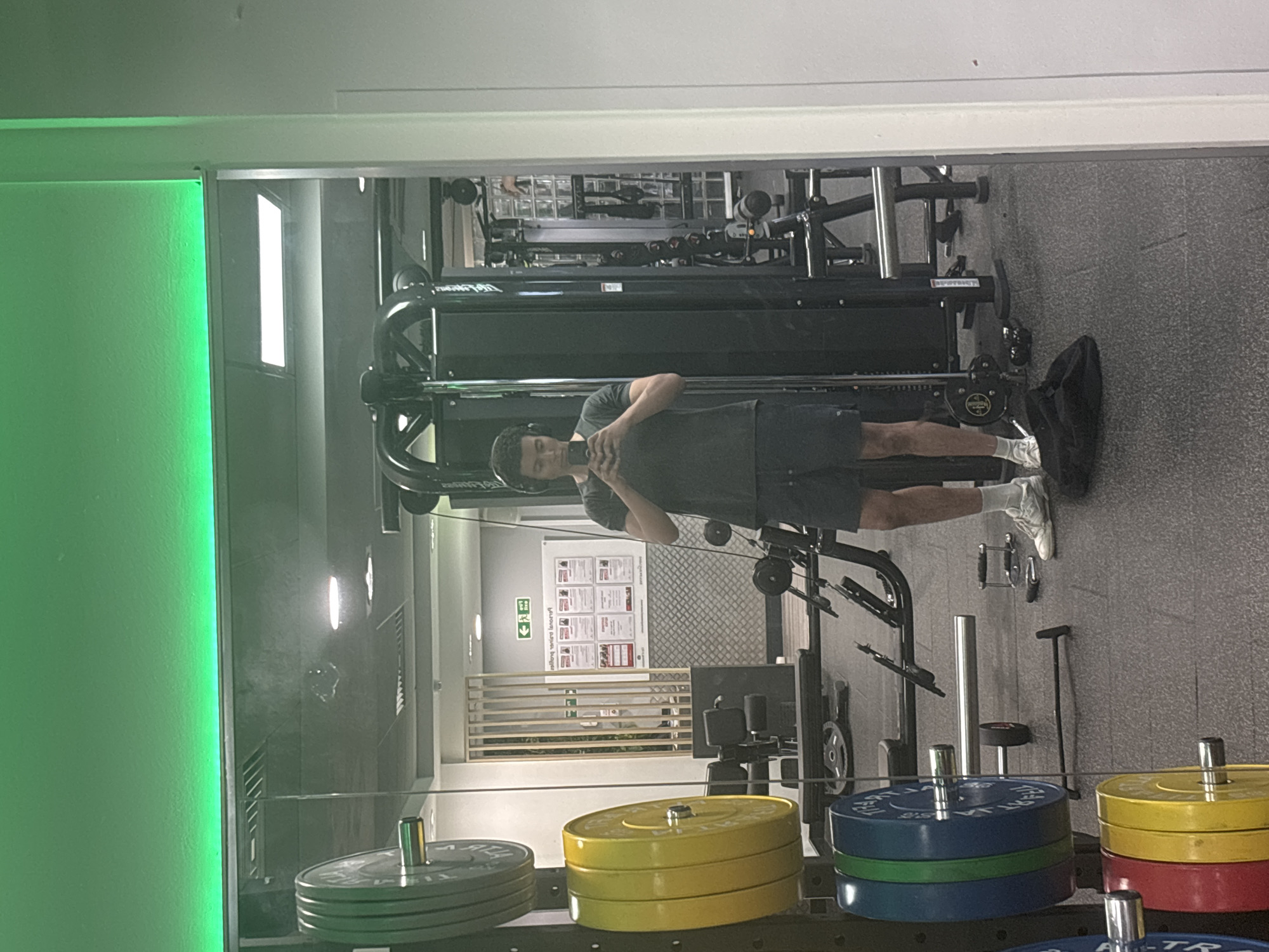 A man stands at a cable machine at a gym, taking a picture of himself in a mirror. The man is wearing a gray t-shirt, dark shorts, and white sneakers. The cable machine is black and has various weights attached. In the foreground are stacks of weight plates in different colors, including green, yellow, and blue. The gym equipment is located indoors. There are safety instructions posted on a nearby wall. Based on the interior decor and gym equipment, the location is likely in London, United Kingdom.