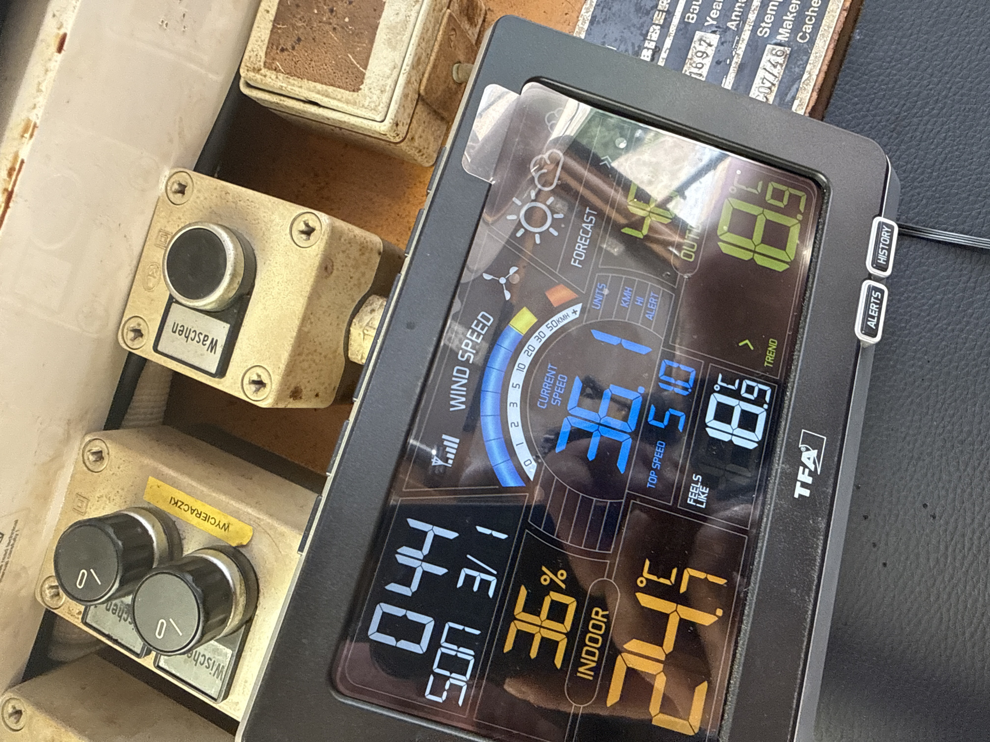 The image shows a close-up of a weather station device with a black casing. The device displays various weather information including temperature, humidity, and wind speed. The time is shown as 0:44, and the day as "SON 3/1". Indoor temperature is 36% and outdoor temperature is 24.7°C. The device also shows wind speed of 36 and a top speed of 51. The forecast icon indicates a partly cloudy day. On the device screen, the TFA logo is visible. In the background, out of focus, are parts of what appear to be older mechanical components, possibly from a boat or a vehicle, with yellow and beige casing and some labels in Polish, including "WYCIERACZKI" which means "wipers". The overall setting suggests an outdoor location, possibly near water, given the context clues from the background objects, which are consistent with the port area of Mikołajki, Poland, a town known for its sailing and marina activities on Lake Mikołajki.