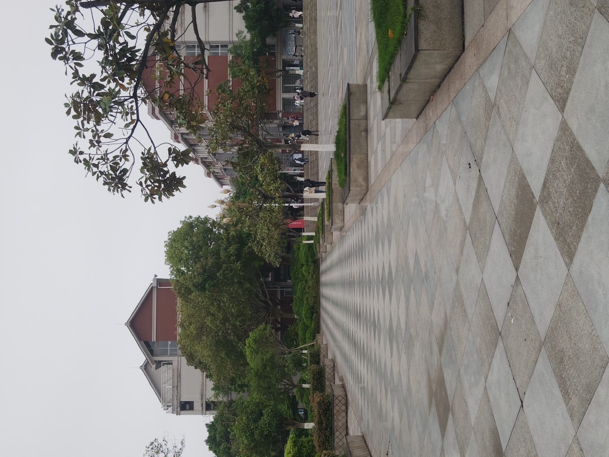 The image depicts an outdoor scene, seemingly a campus or public space within the Chongqing High-tech Zone, China. The foreground is dominated by a checkered tile pathway stretching into the distance. To the left of the path is a strip of greenery with trees and bushes. On either side, the path is bordered by concrete structures. The background features buildings, one with red brick accents and another with a white facade. Several people are faintly visible in the background, suggesting a moderately populated area. The sky appears overcast and grey.