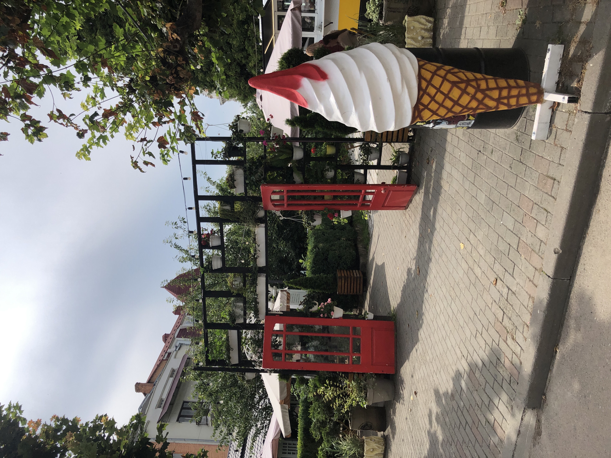 The image depicts an outdoor scene in Kolomyia, Ukraine, likely a cafe or restaurant entrance. In the foreground, a large, whimsical sculpture of a vanilla ice cream cone with red syrup drips stands on a metal base next to a black barrel. The ground is paved with light brown bricks.
To the left, a bright red, two-doored gate with glass panes is open, leading into a courtyard or garden area. Behind the gate, a black metal structure is adorned with numerous potted plants, including greenery and some flowering plants.
The background reveals a street with parked cars and buildings, one with a red-tiled roof and chimneys. To the right, a person is partially visible near a food truck or stall with a beige awning and a yellow lower section. More potted plants and greenery are scattered throughout the scene, creating a welcoming, decorative atmosphere. The sky is overcast.