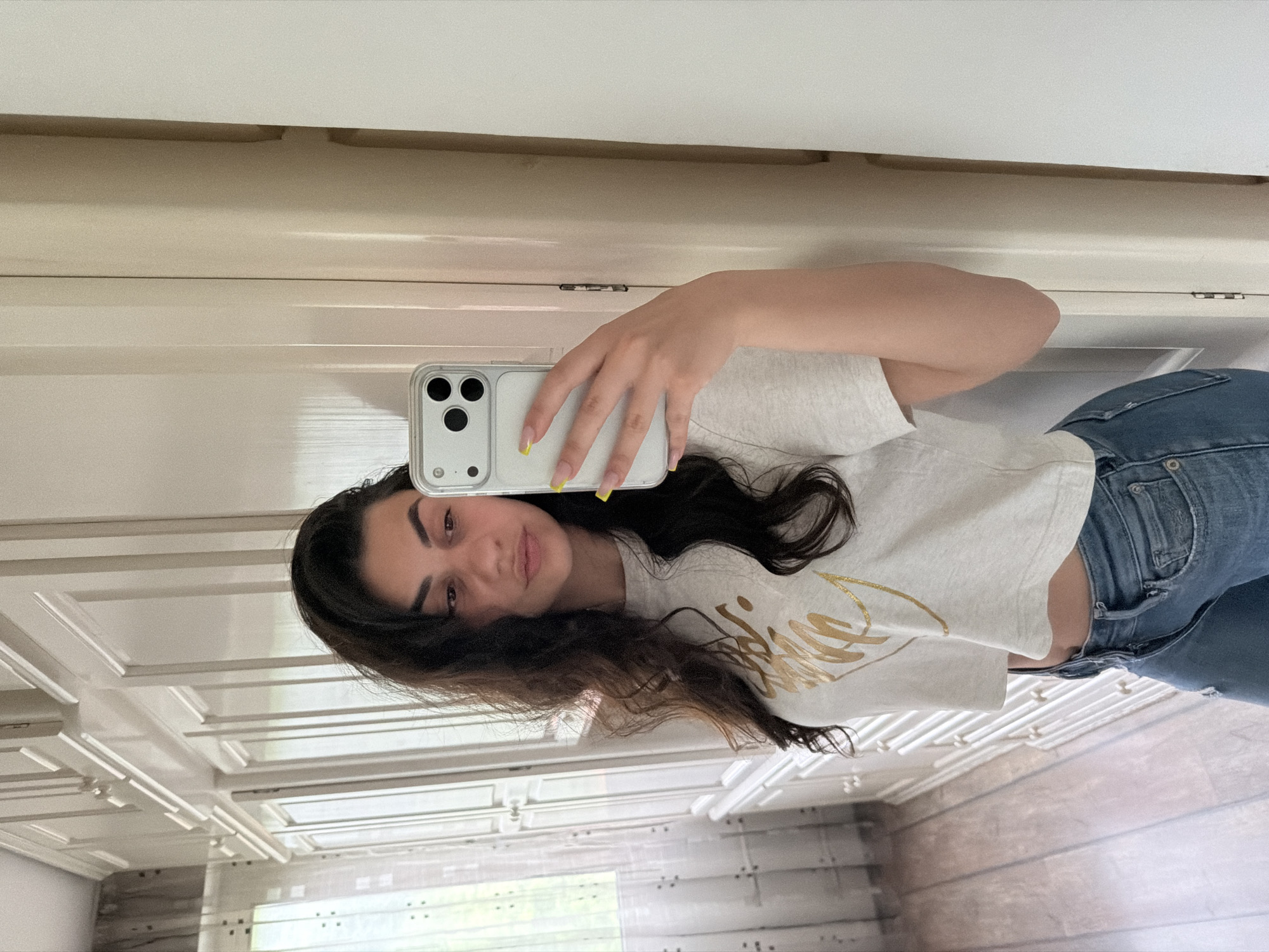This image captures a young woman taking a mirror selfie indoors in Gabrovo, Bulgaria. The photo is oriented vertically, showing the woman from the chest down, holding a white smartphone with a transparent case in her left hand. Her nails are painted with vibrant yellow tips.
The subject has long, dark, wavy hair that flows over her shoulders. She is wearing a light-colored, possibly off-white or light grey, short-sleeved crop top adorned with a stylized gold design or text on the front, which is partially obscured and not clearly decipherable. She is also wearing blue jeans. Her facial expression is calm and neutral, with a slight, soft smile as she looks directly towards the phone's camera.
The background, visible as a reflection in the mirror, features light-colored interior elements that appear to be white or off-white cabinetry or decorative wall panels with horizontal and rectangular moldings. In the lower left portion of the reflected background, a glimpse of outdoor greenery through a window suggests it is daytime. The setting appears to be a well-lit domestic space, possibly a kitchen or bathroom.