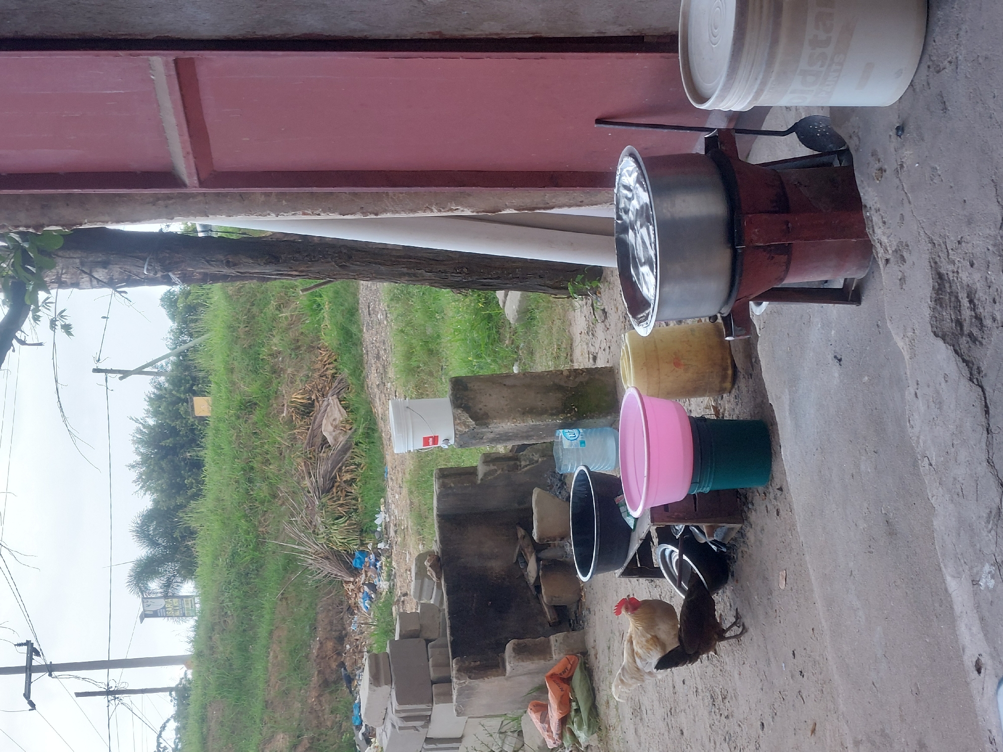 A scene in Dar es-Salaam, Tanzania, displays a rustic cooking setup outdoors. A large metal pot sits on a wood-fired stove constructed from cement blocks. Various plastic containers, including a pink bowl, a green bucket, and a blue water bottle, are arranged nearby. A white bucket with printed text is also visible. Chickens roam near the stove and containers. In the background, there is a green field, trees, and power lines. The scene is captured from a slightly elevated viewpoint, showing the surrounding environment. The image appears to be taken during the day under overcast conditions.