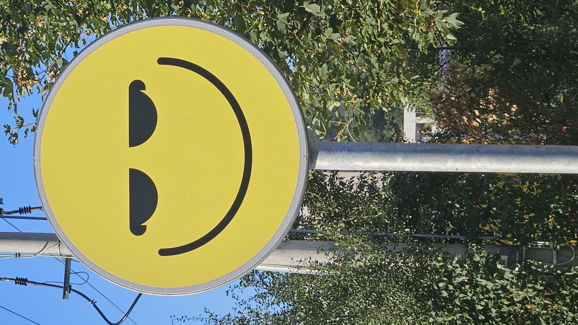A yellow circular sign with a stylized smiley face is mounted on a metal pole. The smiley face has black eyes, depicted as angled ovals, and a curved black line for a mouth. The sign has a reflective silver border. The background consists of green trees, foliage, and a clear blue sky. A utility pole with electrical wires is visible to the left of the smiley face sign. In the distance, a white building with windows can be seen through the trees. This sign appears to be located in the city of Prešov, Slovakia, as indicated by the context provided. The scene depicts an outdoor environment, likely a public area.
