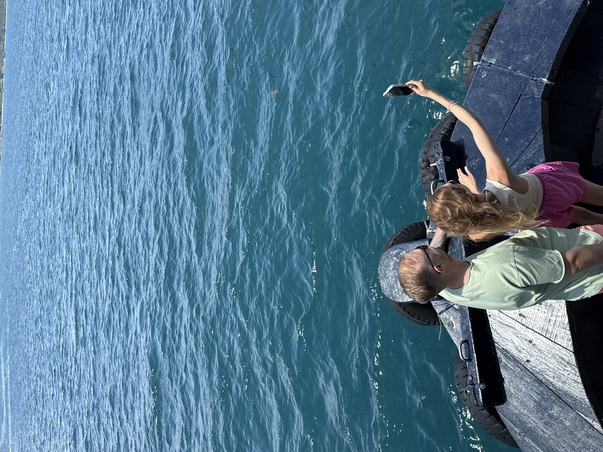 Two individuals, a male and a female, stand on what appears to be a pier or a dock. The female is holding a smartphone and appears to be taking a picture. The water is a deep turquoise, slightly rippled. The location is Bac Nha Trang Ward, Vietnam.
