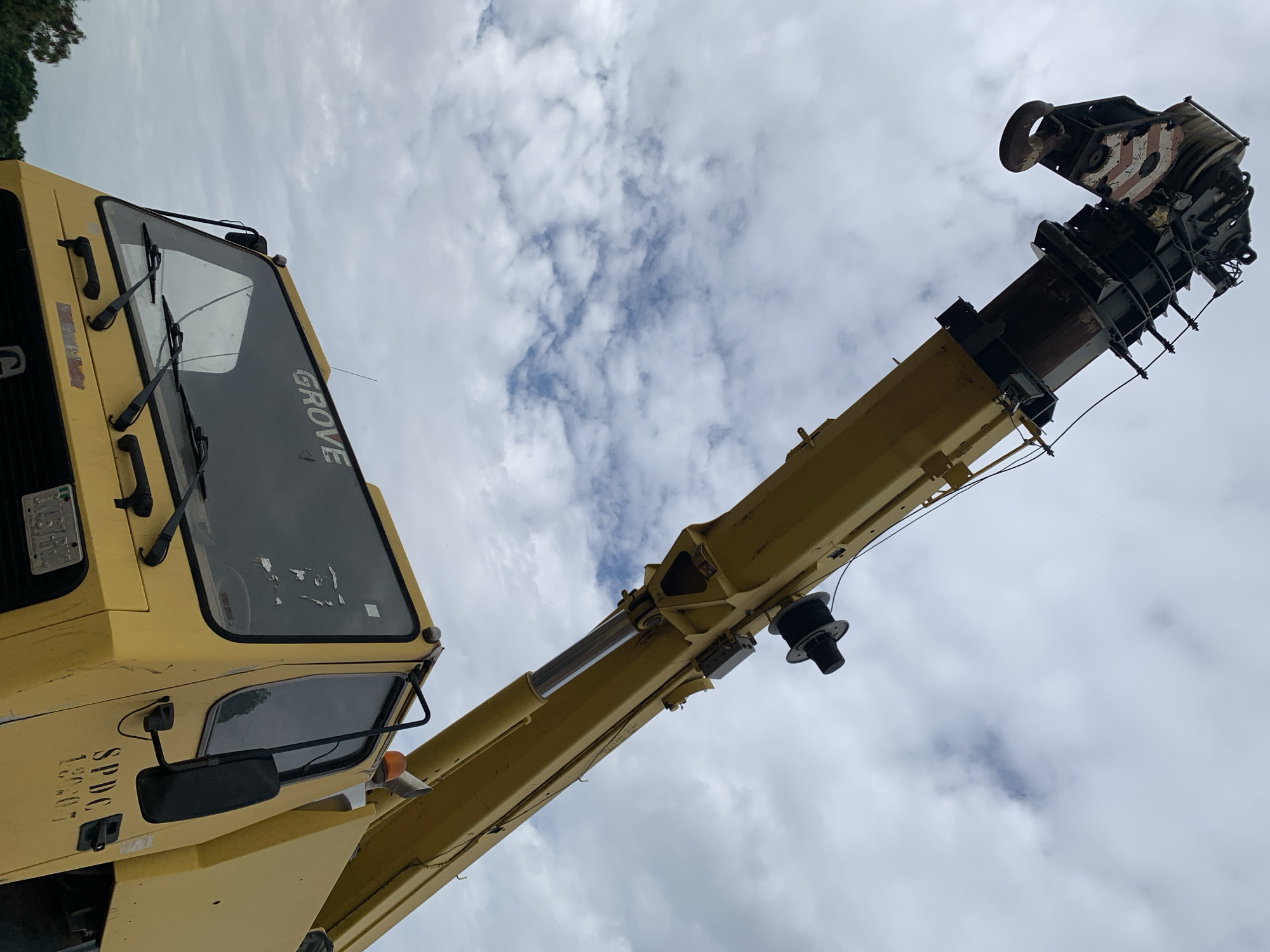 A yellow mobile crane is depicted from a low-angle perspective against an overcast sky with scattered clouds. The crane's main boom extends diagonally upwards from the lower right towards the upper left. Attached to the boom's end is a multi-sheave hook block, prominently marked with white and red diagonal safety stripes. A hydraulic cylinder is visibly extended, supporting the boom. The crane's cab, also yellow, occupies the lower left portion of the frame, featuring a large windshield. The brand name "GROVE" is visible on the upper part of the windshield. On the side of the cab, the alphanumeric designation "SPDC 130407" is present. A Nigerian vehicle license plate is partially visible at the front of the cab, displaying the characters "DL4GYPHG" and the word "NIGERIA" below. The background consists primarily of a light grey, cloudy sky with hints of blue in gaps between clouds. Minimal dark green foliage is discernible in the extreme lower left corner. No individuals are present in the scene. While the visible license plate and the "SPDC" marking indicate a Nigerian operational context, the specific location in the city of Rafin Par is not verifiable from the visual information within the image.