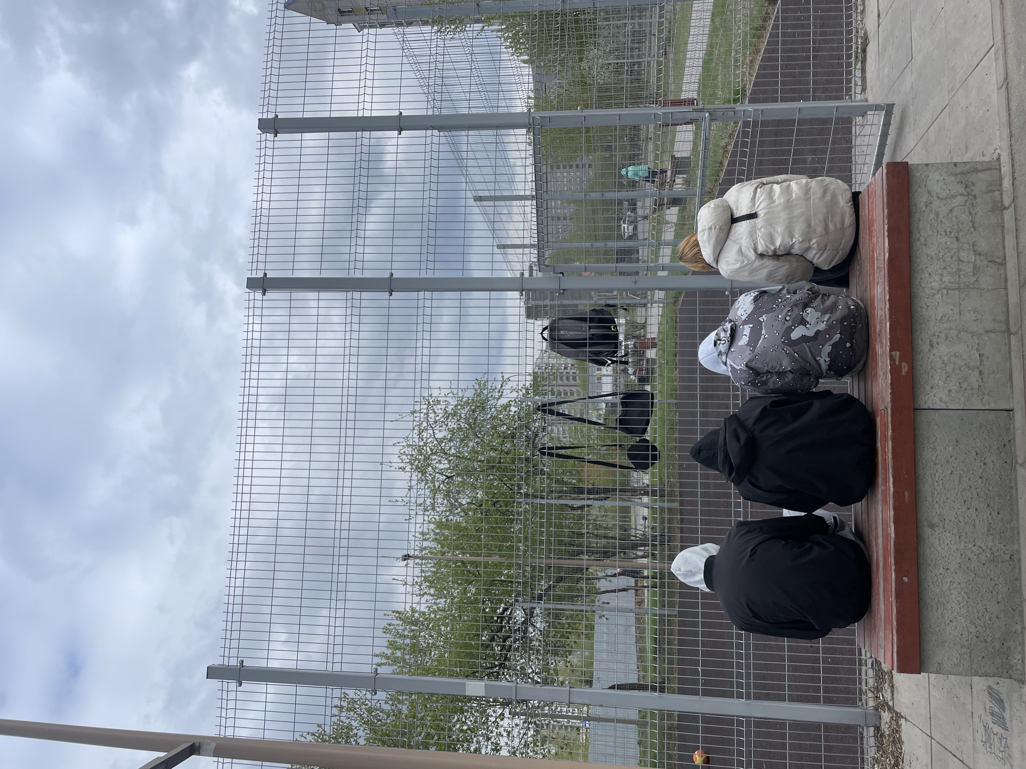 Four individuals are sitting on a red bench, facing away from the camera and towards a wire fence. Beyond the fence, there is a tree with sparse green leaves and a playing field with a dark surface. Buildings are visible in the background, suggesting an urban environment. Backpacks are hanging on the fence. The sky is overcast. The location is most likely Krasnoyarsk, Russia.