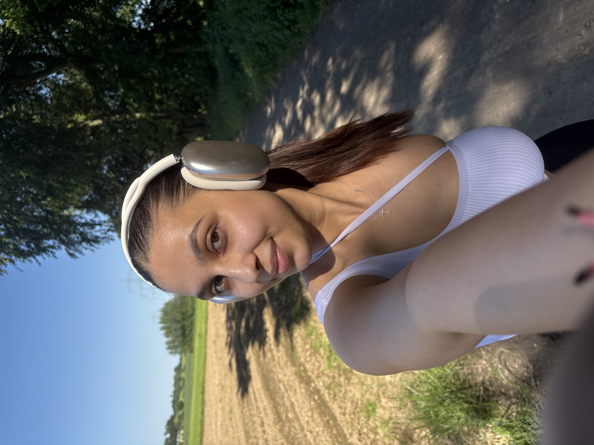 The image shows a young woman in a white tank top, wearing large headphones, possibly Apple AirPods Max. She is taking a selfie. There is a faint tattoo visible near her shoulder. The background suggests a rural or semi-rural setting. To the left, the sky is visible, indicating a clear day. A field is visible, some parts appearing freshly harvested or dry. Trees line the horizon. The photograph's location can be identified by the presence of fields and open skies, in addition to being set on a path for walking, which are often found on the outskirts or near green spaces of the city. Given the information that the location is Viersen, Germany, the photo could have been taken near one of the agricultural areas or parks on the periphery of the city.