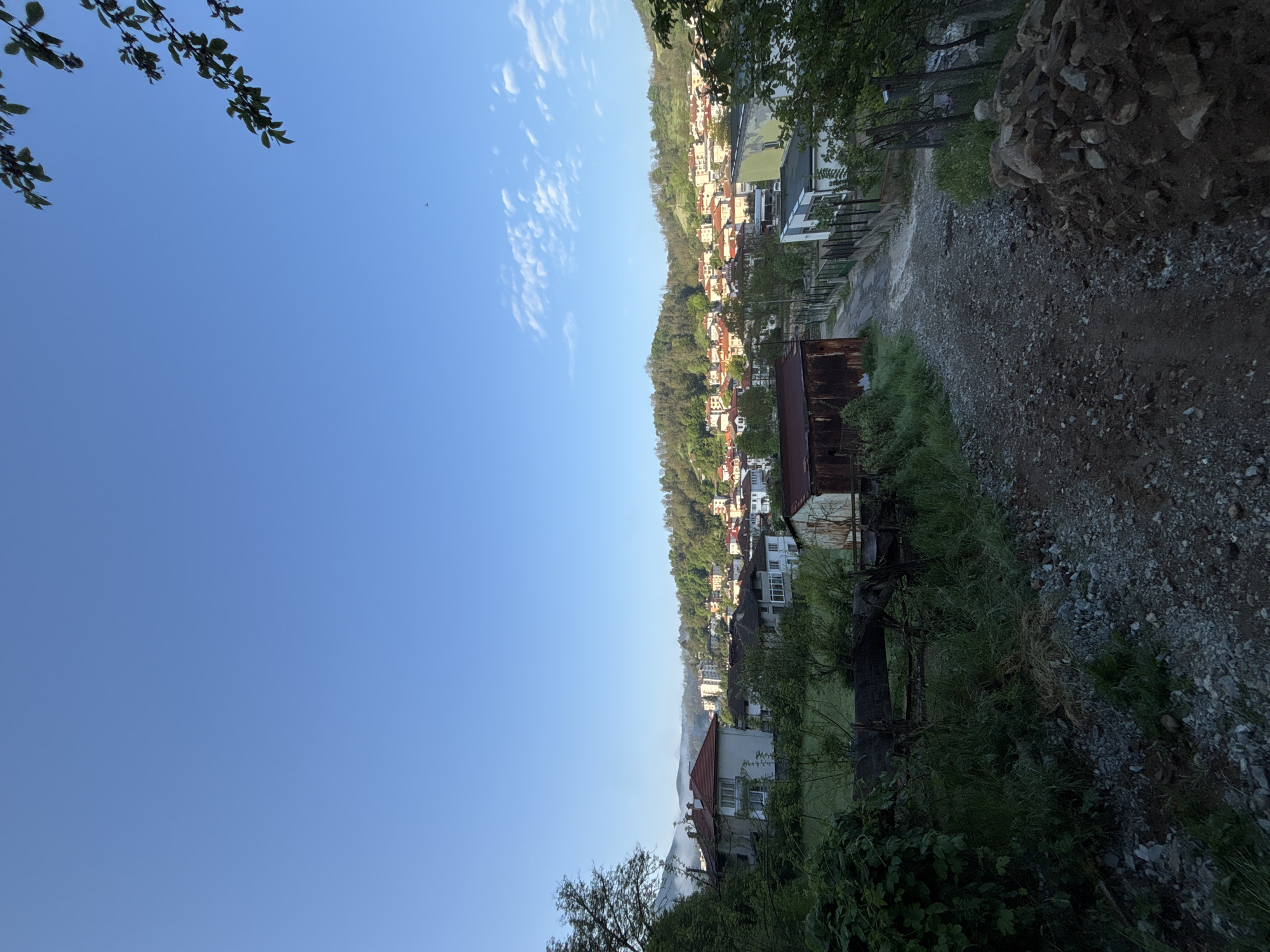 The image shows a view of Zlatograd, Bulgaria, from an elevated position. The camera is oriented approximately 90 degrees counter-clockwise. In the foreground, there's a dirt road or path with vegetation along its edge. To the right is a pile of rocks. Further into the middle ground are residential buildings clustered along the hillside. A clear blue sky occupies a significant portion of the frame. There are small, puffy clouds dispersed in the upper center. A few tree branches are visible in the upper left. A bird flies in the middle left of the image.
